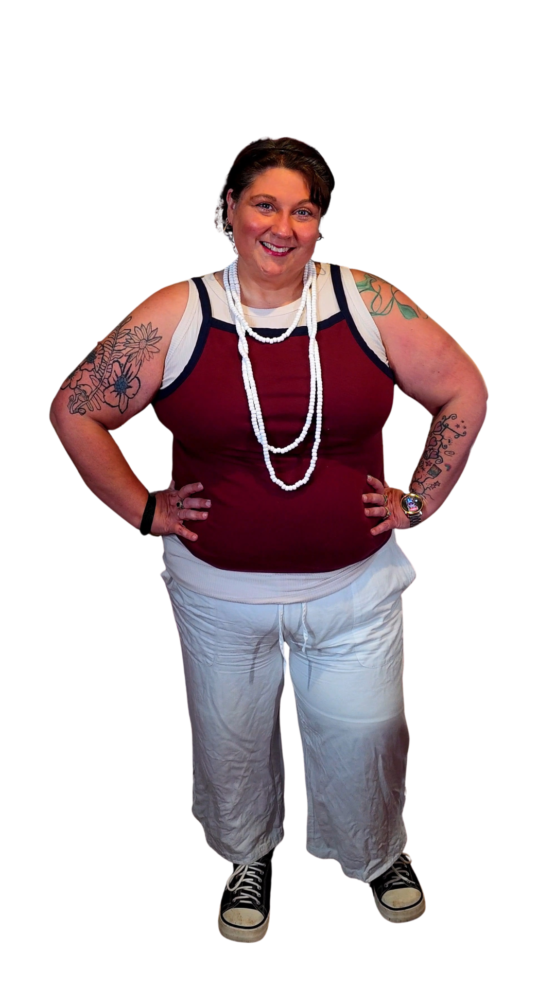 A woman standing with her hands on her hips, smiling, wearing a sleeveless maroon top with white and blue accents, white pants, black sneakers, layered necklaces, and has tattoos on both arms.