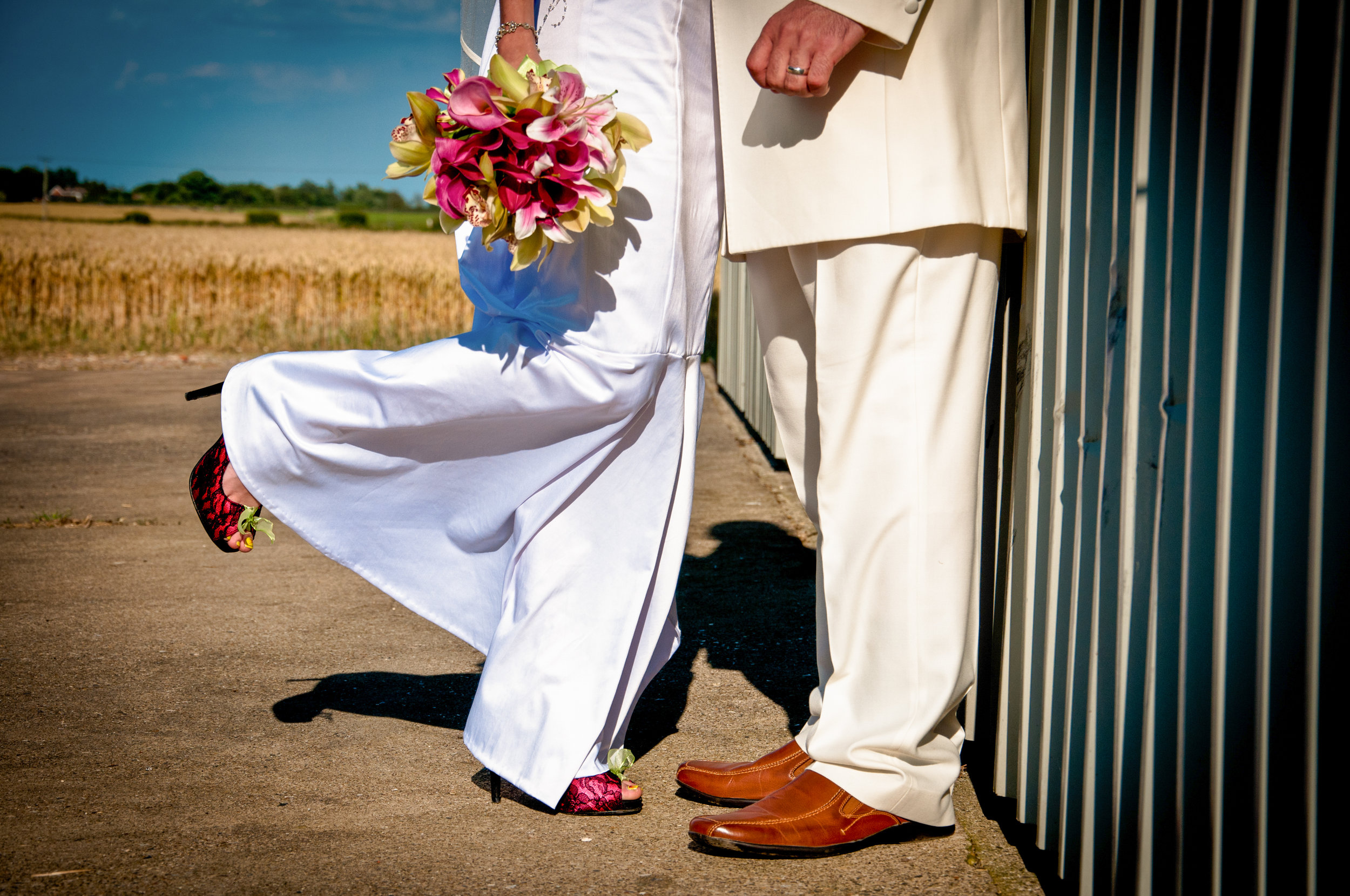 A bride and groom lean against a shed in a brilliant blue skied day, the bride is leaning against the groom her leg popped up. The bride is holding flowers that are vibrant pink and the groom is in a cream suit.