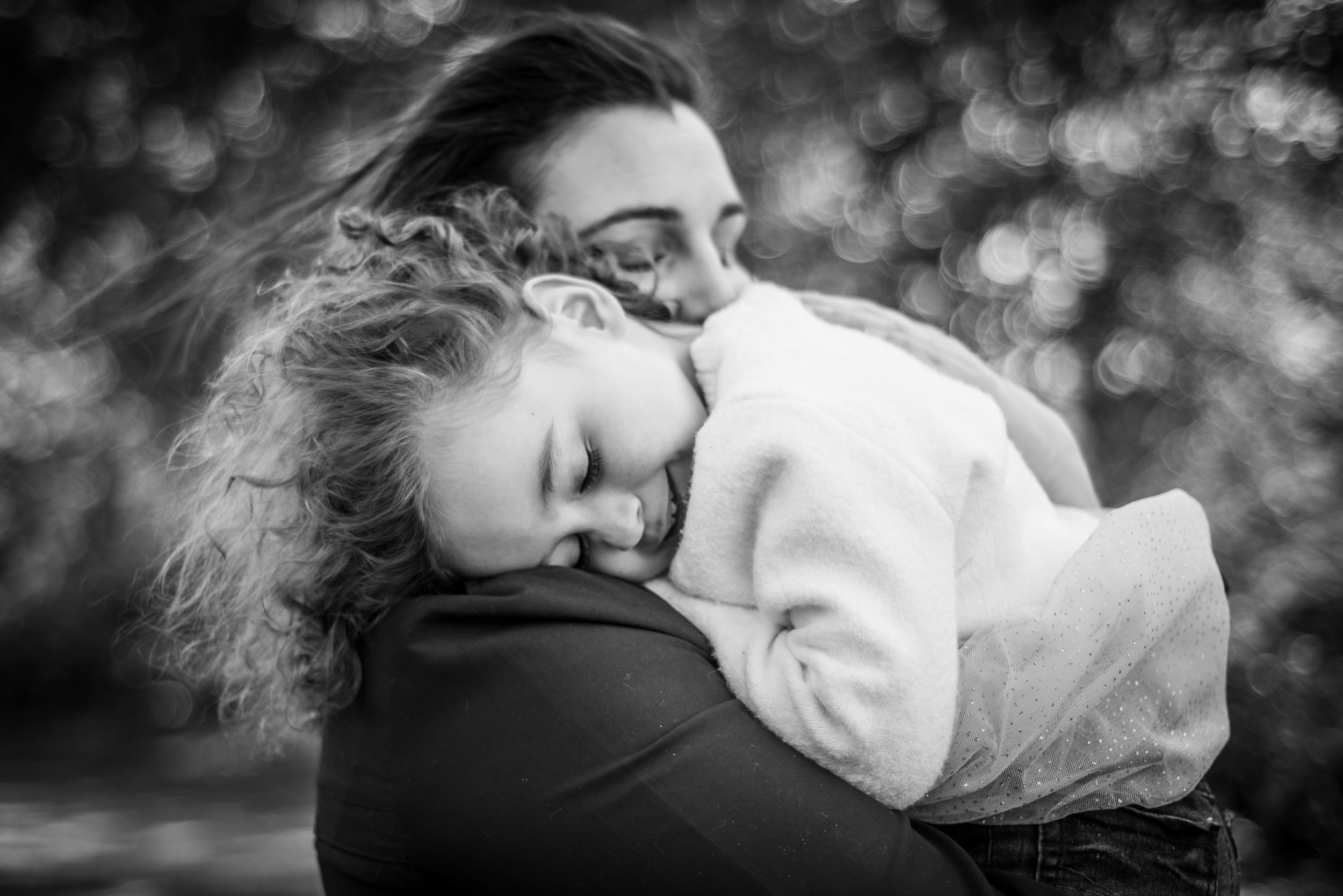 A mother embracing her duaghter on Chorelywood Common, bracing against the wind. The daughter has a slight smile on her face while the mother’s eyes are closed and her expression is loving.