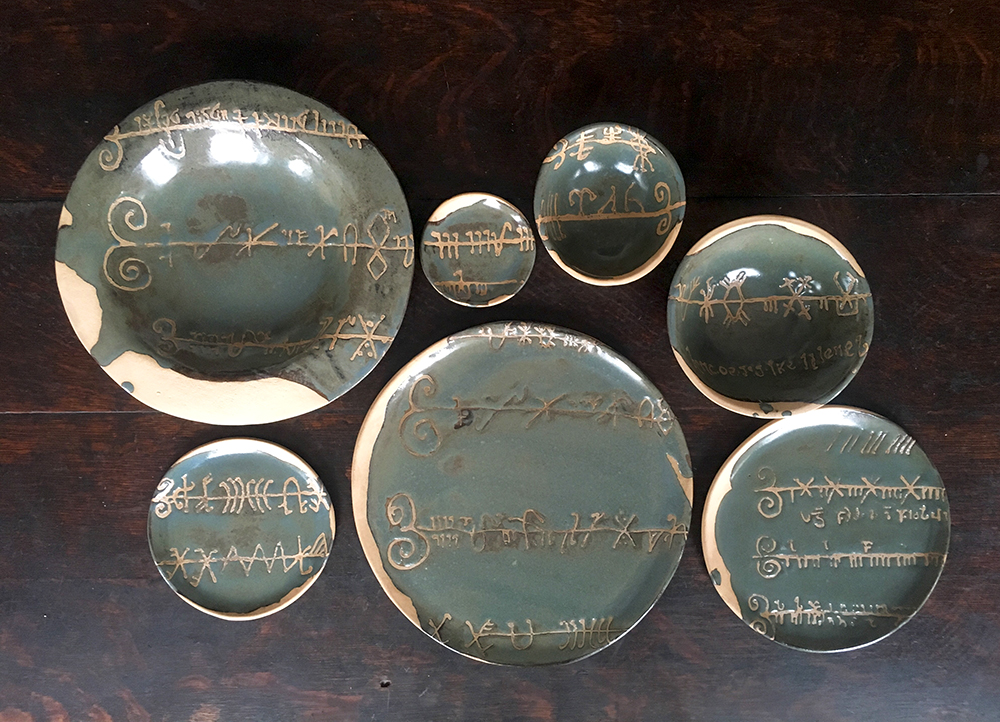 ancient celitc ogham script that resembles a music score across place setting including charger, dinner, 7”, 5” plates, medium, low & small bowls in moss on blush clay