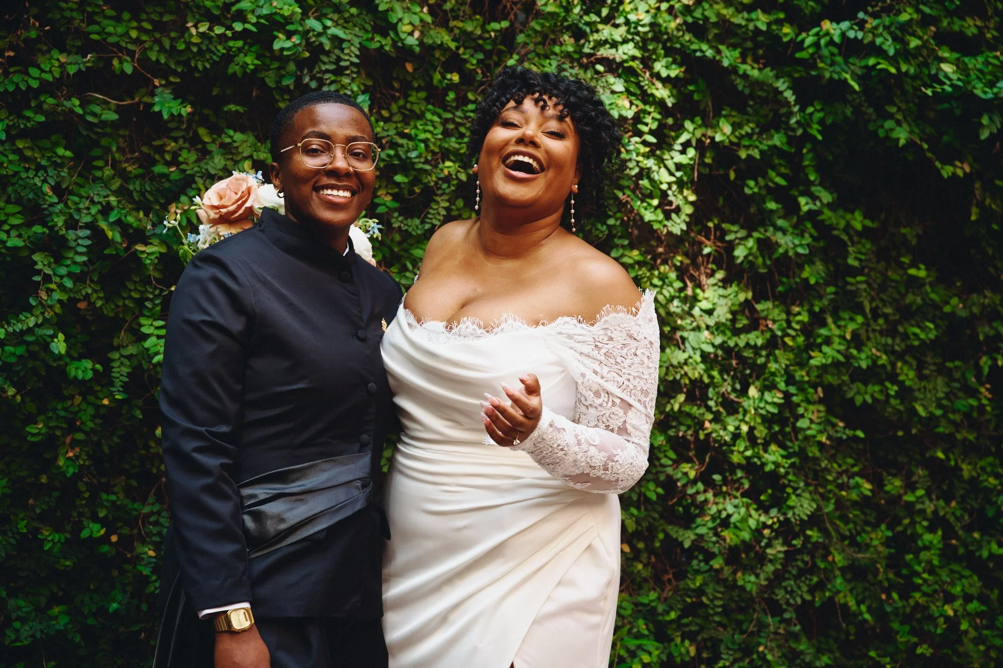  A black and white portrait of a couple in front of a wall of greenery 
