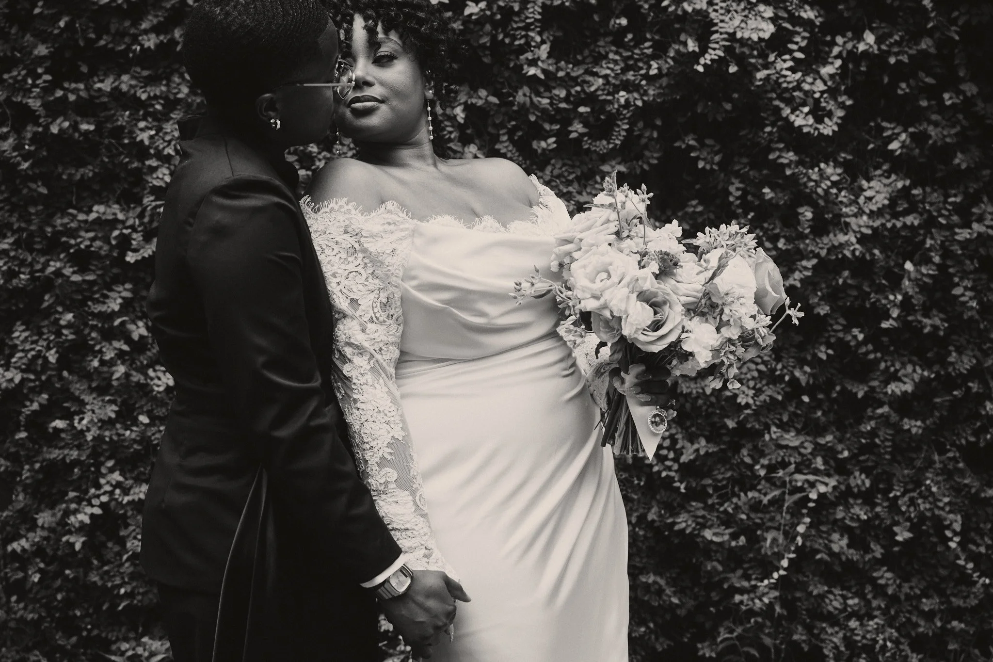  A black and white portrait of a couple in front of a wall of greenery 
