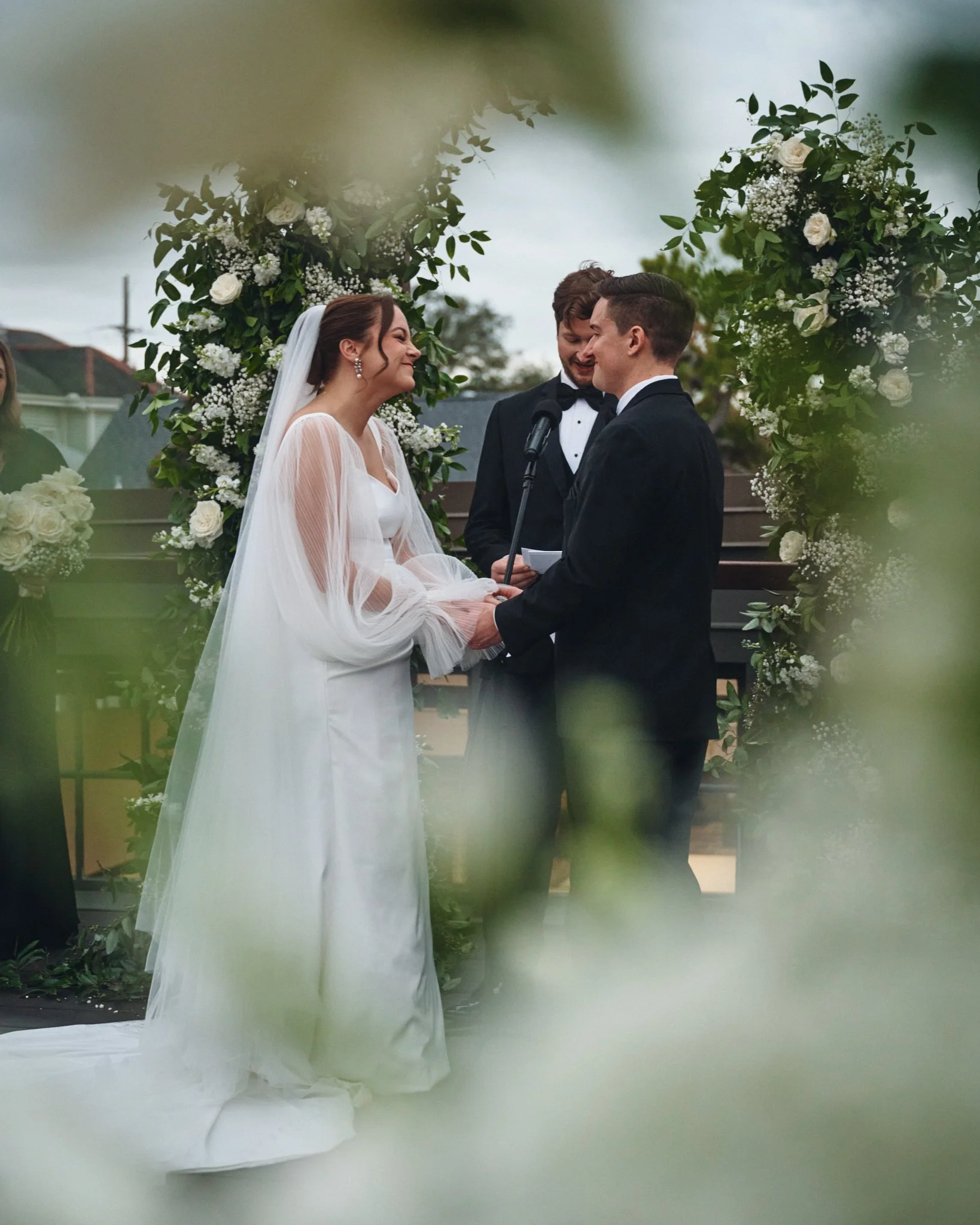  A bride and groom holding hands during their vows, framed through white roses at Capulet in New Orleans 
