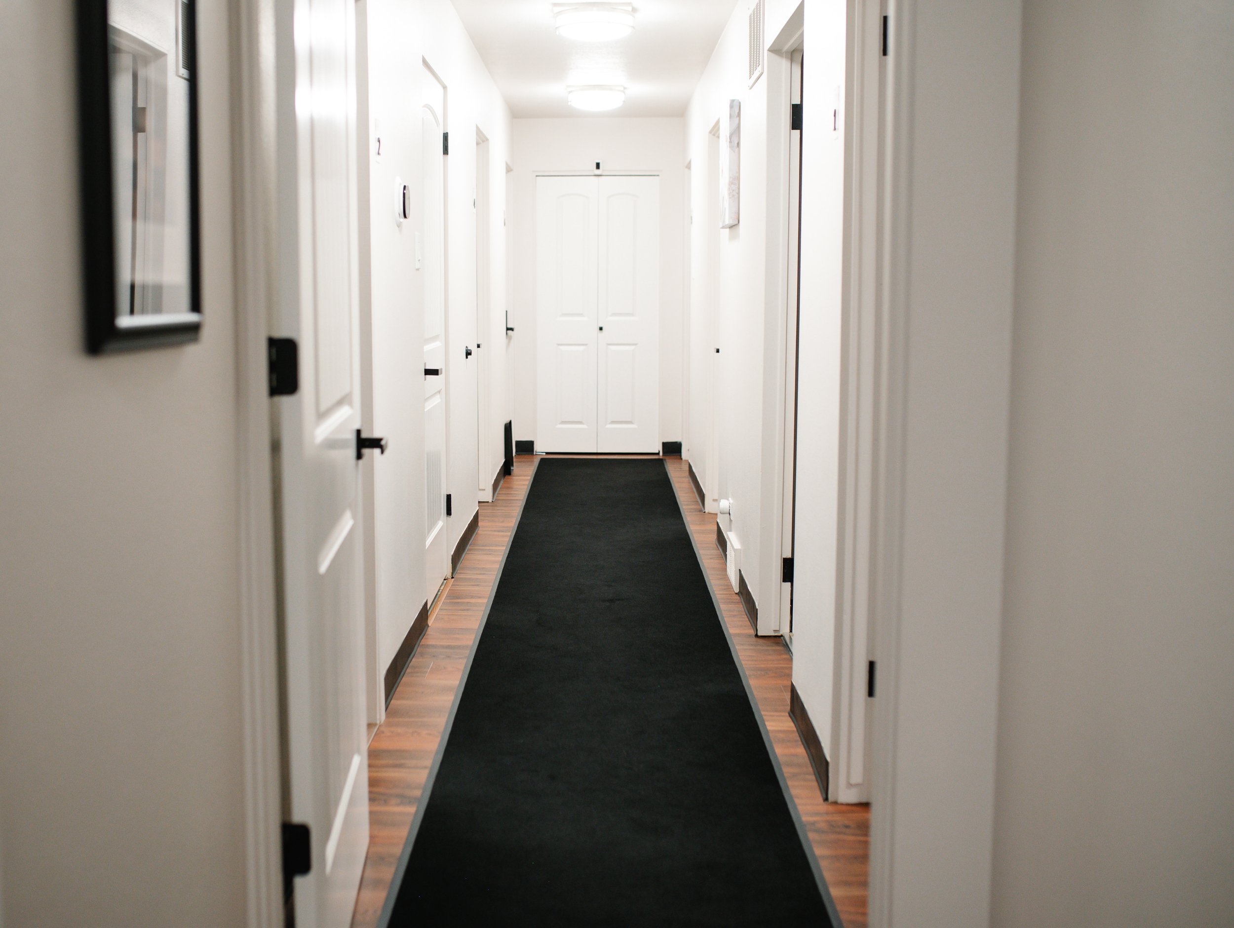 A hallway with white walls, wooden flooring, black baseboards, closed white doors on both sides, and a black rug running down the center. Fluorescent ceiling lights illuminate the corridor.