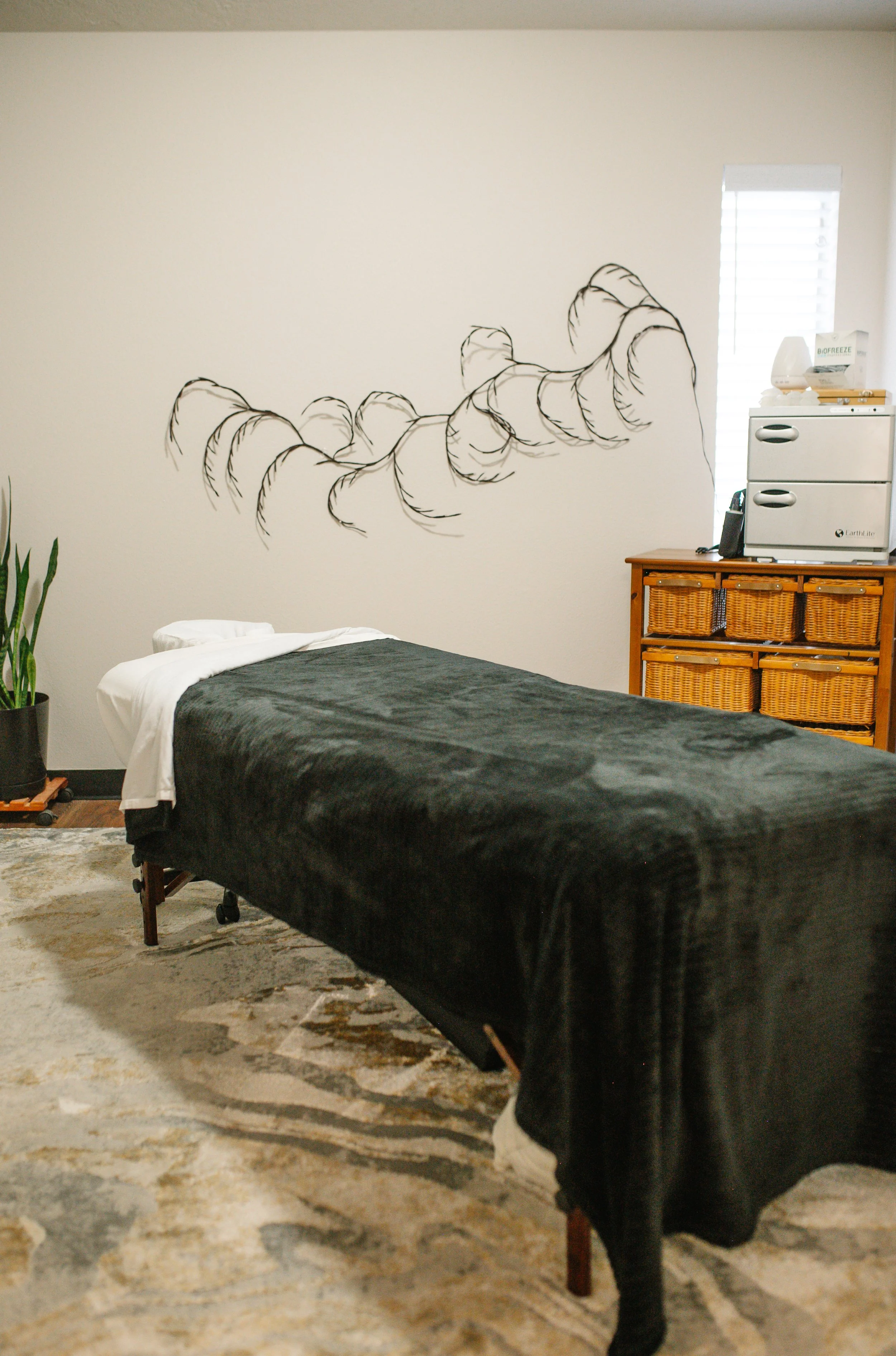 A spa or massage room with a massage table covered with a black towel, a white sheet at the head, and a patterned rug underneath. To the side, there is a tall green plant and a small wooden cabinet with storage baskets. On top of the cabinet, there is a massage machine and some other items. The wall has a black line drawing of a caterpillar or worm. There is a window with blinds, allowing natural light into the room.