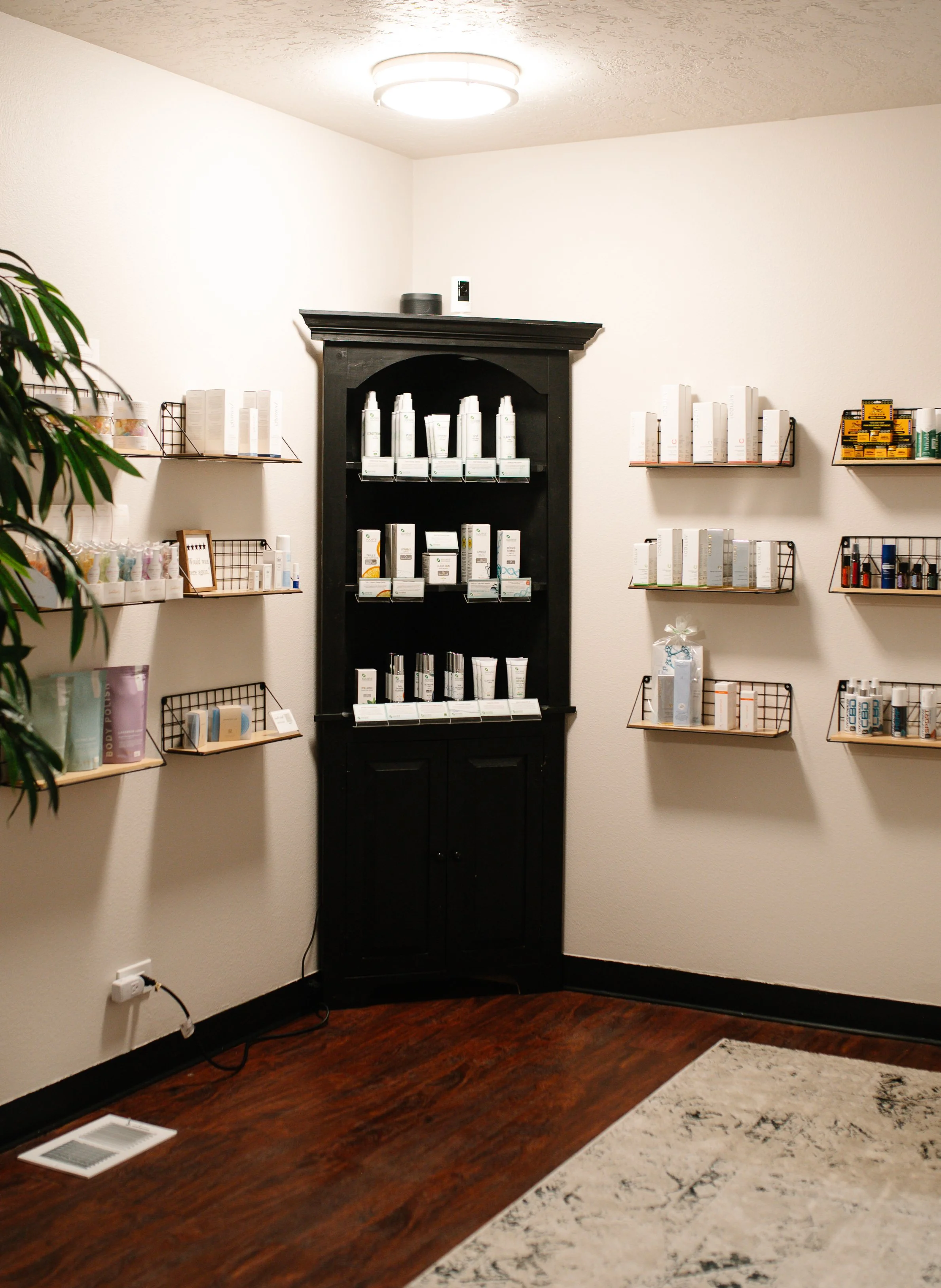 A room with cream walls, a wooden floor, and a patterned area rug. Shelves with various skincare or cosmetic products are mounted on the walls. A black cabinet displays more products, and there is a ceiling light fixture.