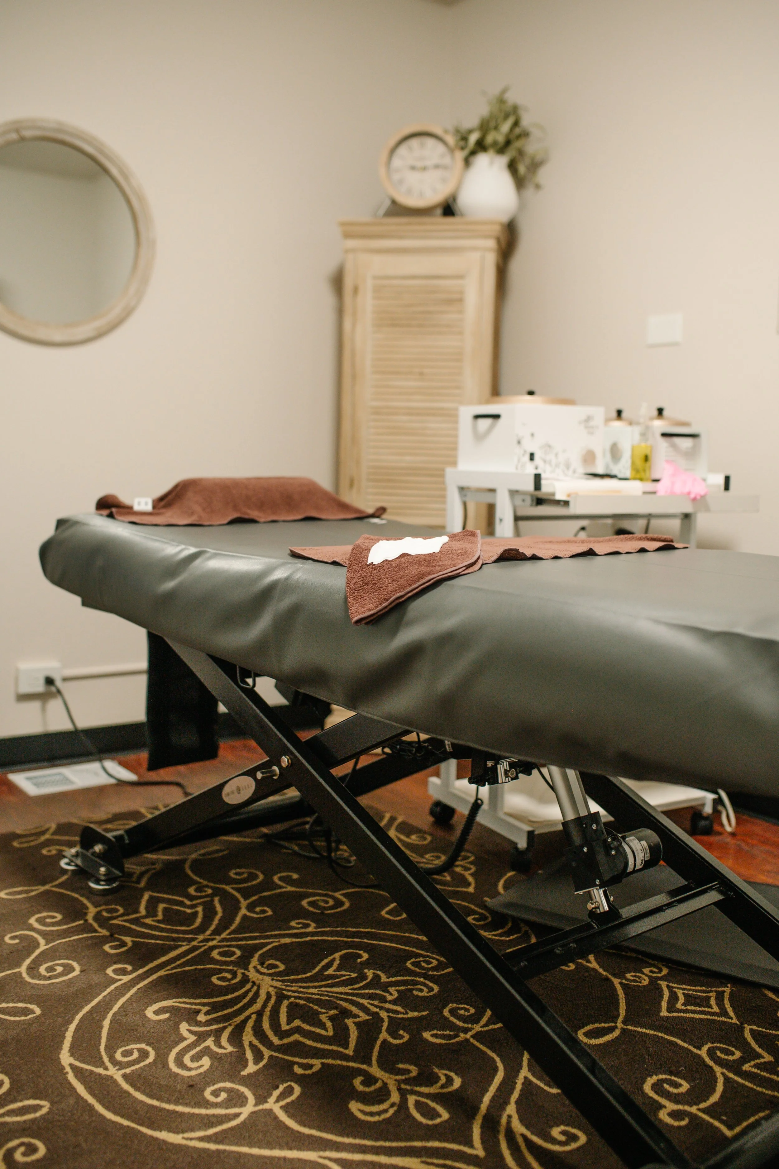 Medical examination room with an adjustable massage table covered with a gray sheet, a brown towel, a decorative rug with yellow and brown patterns, a wooden cabinet with a clock and a white vase with green foliage, and a table with medical supplies.