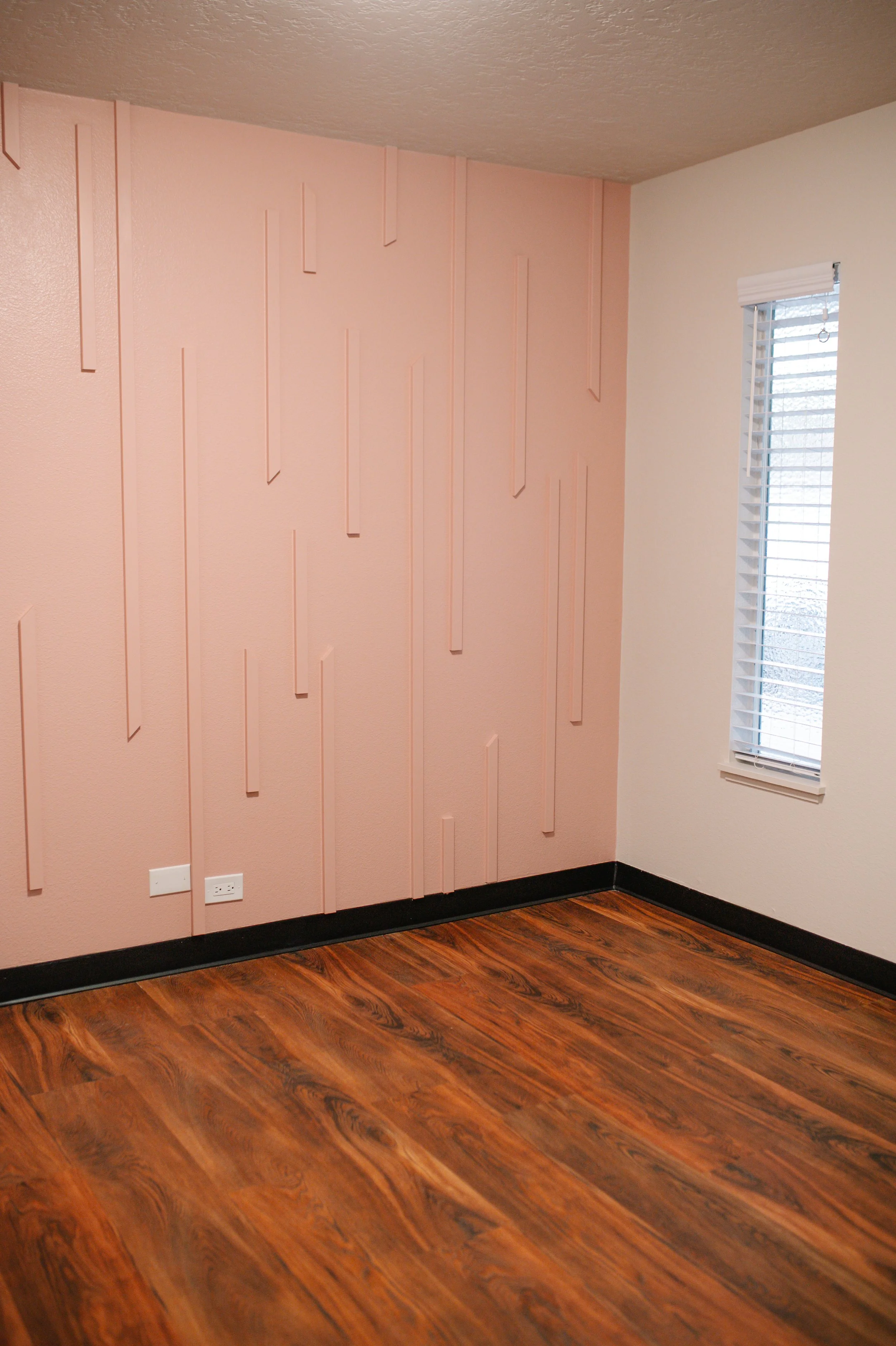 An empty room with pink textured walls, a window with blinds, and wooden flooring.
