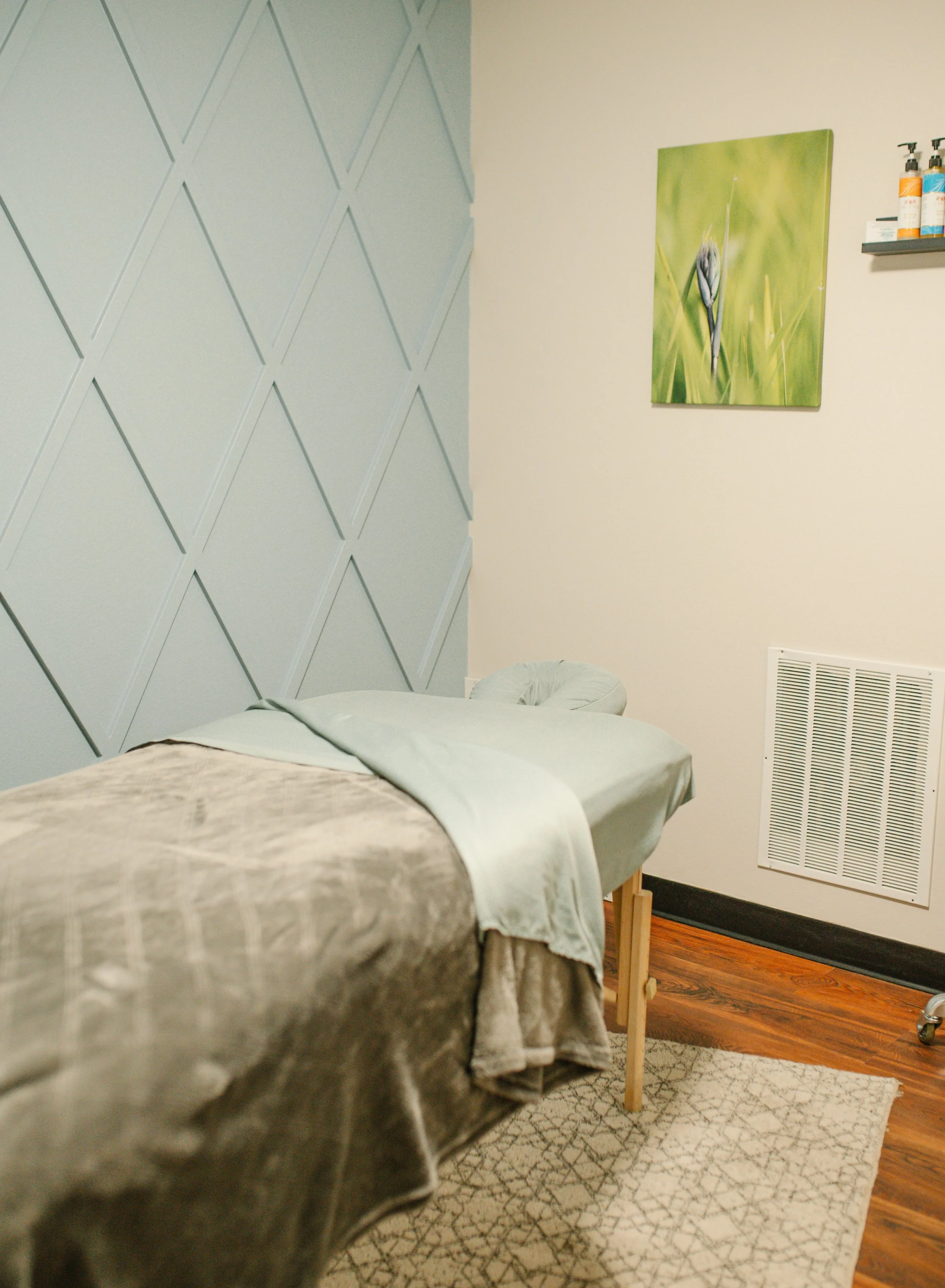 A massage table covered with beige linens in a spa room, with a framed botanical photograph on the wall and a small shelf with bottles of lotion or oil.