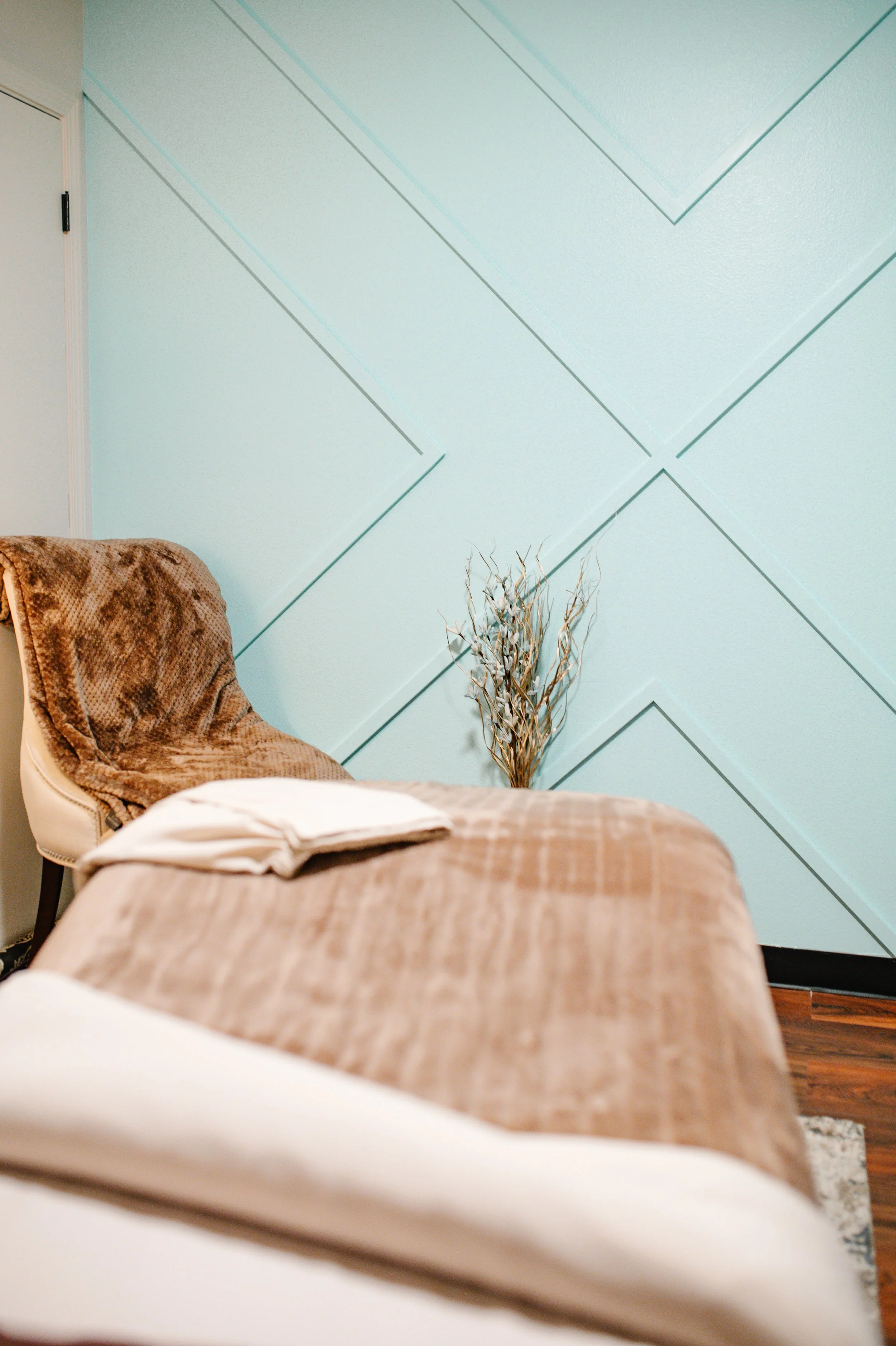 Part of a room with a mauve-colored bed, a chair with a brown blanket, a wall with decorative blue-green panels, a decorative dried plant, and a closed door.