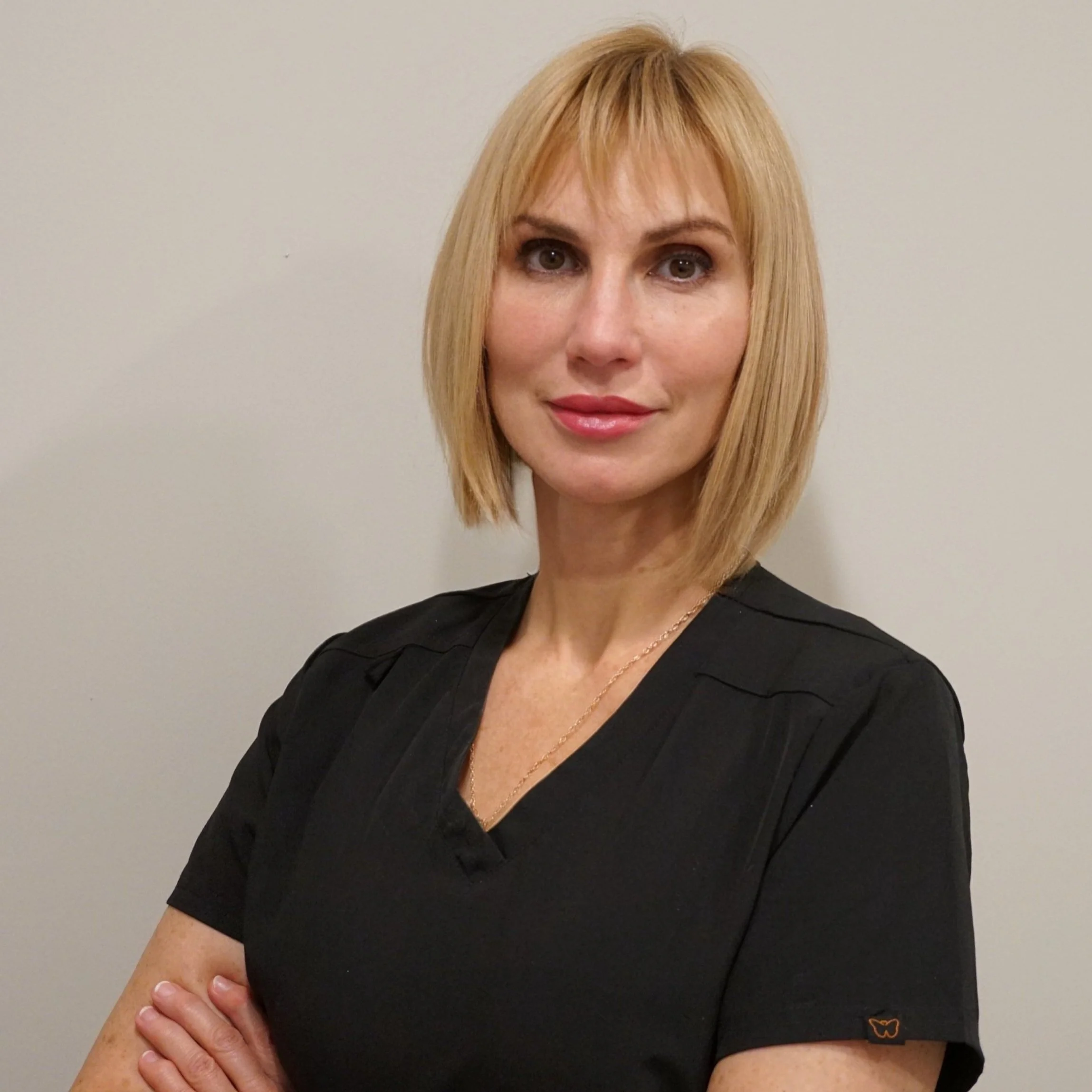 A woman with short blonde hair wearing a black scrub uniform, standing with arms crossed against a plain background.