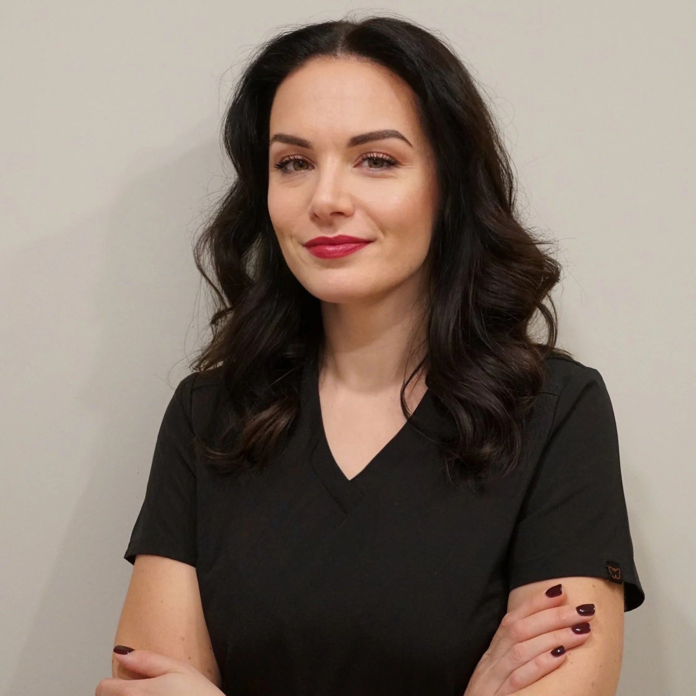 A woman with dark hair wearing a black top and standing with arms crossed, against a plain background.
