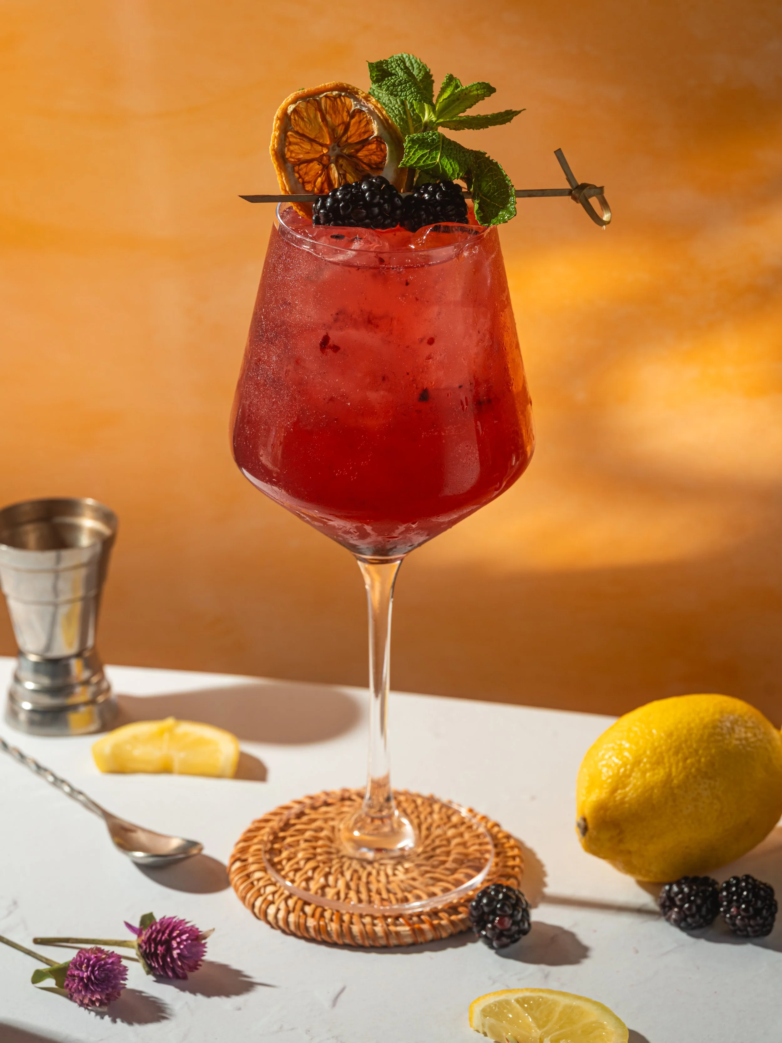 A pink cocktail in a tall wine glass garnished with mint leaves, blackberries, and a slice of dried citrus, placed on a wooden board with additional blackberries and mint leaves around it. There is a small metal container with more blackberries, a spoon, and a light-colored background.