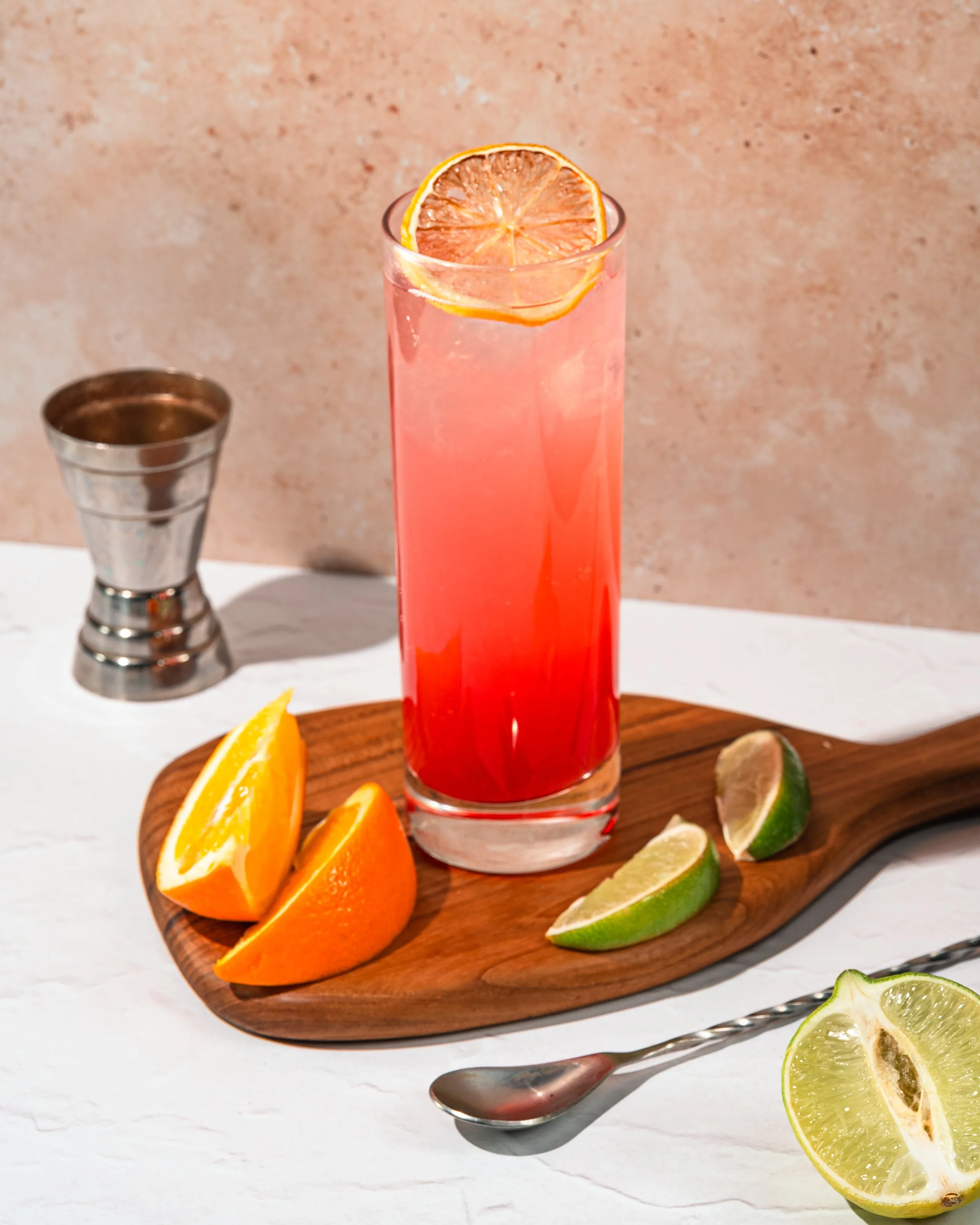 Tall glass of pink citrus drink garnished with a dried orange slice, placed on a wooden board with orange, lemon, lime wedges, a spoon, and a metal jigger on a white table with a textured beige background.