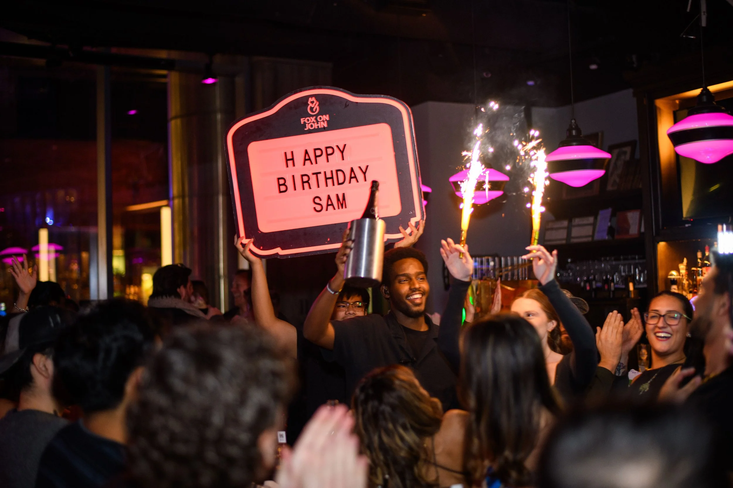 People celebrating at a birthday party, some holding sparklers and a sign that reads 'HAPPY BIRTHDAY SAM', in a lively indoor setting with colorful lighting.