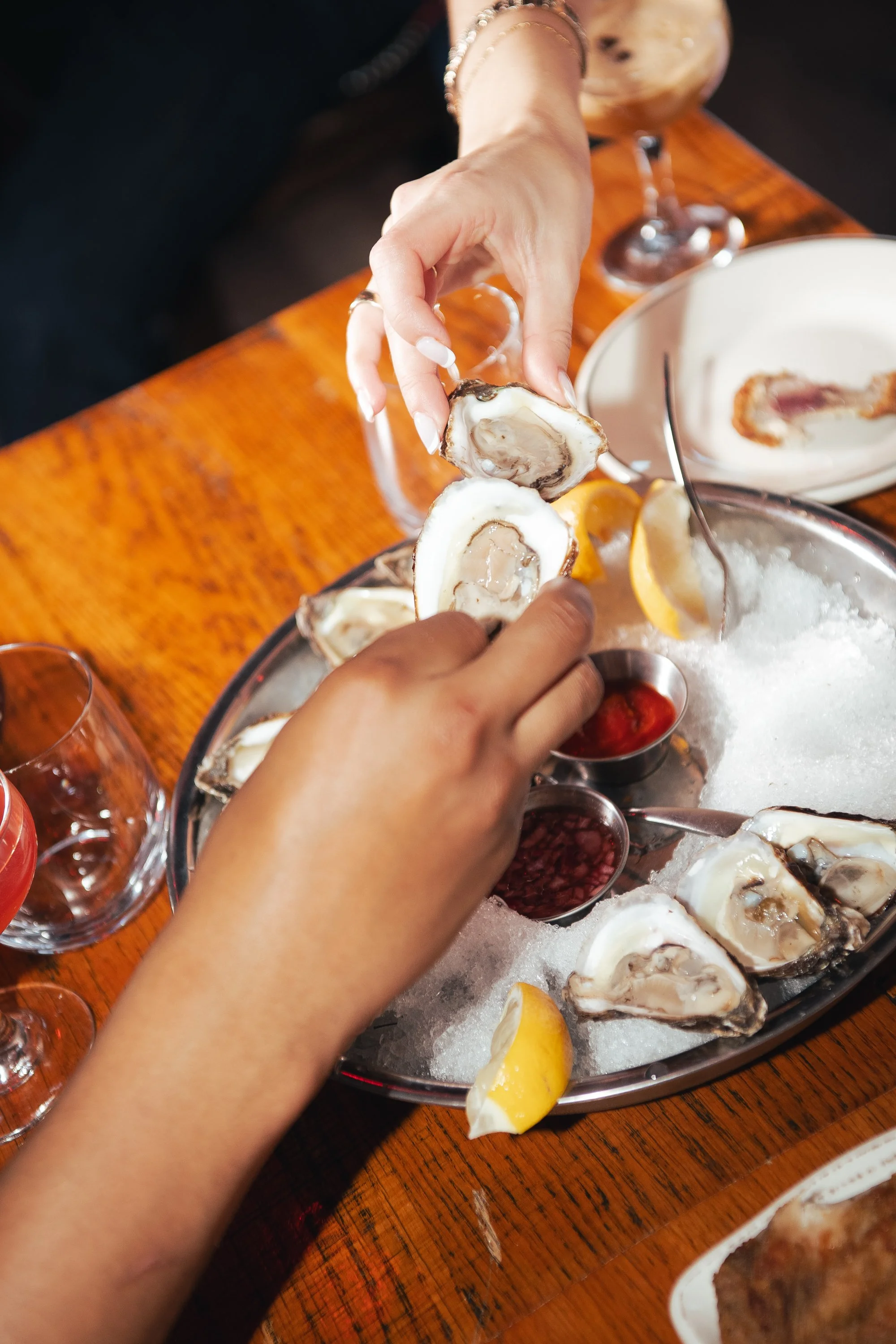 A seafood platter with raw oysters on the half shell served on a bed of ice, garnished with lemon wedges, with two small bowls of cocktail sauce and mignonette in the center, placed on a wooden table. Two cocktails in martini glasses are in the background and a blue book is visible behind the drinks.