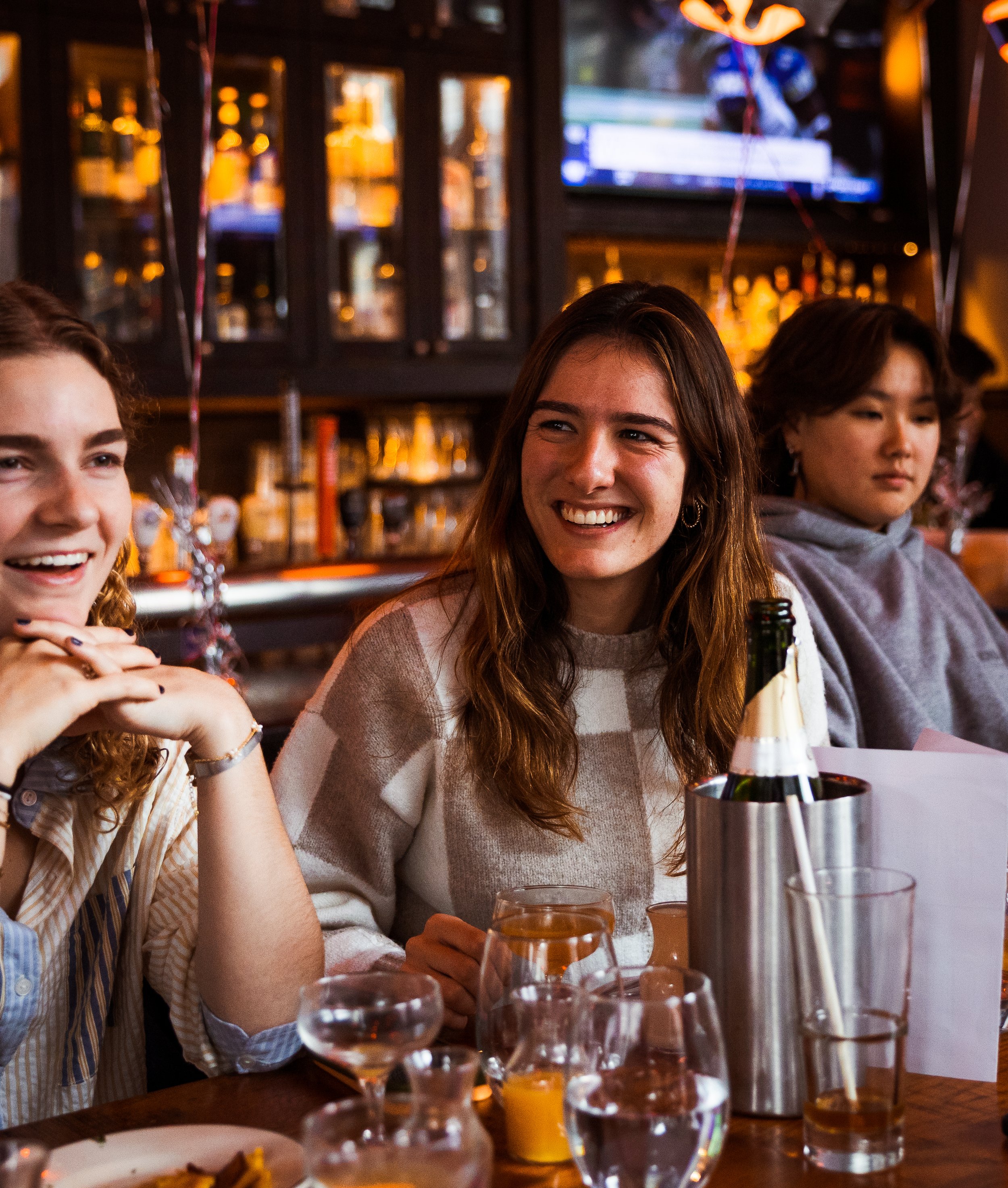 Three young women enjoying a celebration at a bar or restaurant, with drinks and a bottle of champagne on the table, smiling and conversing.