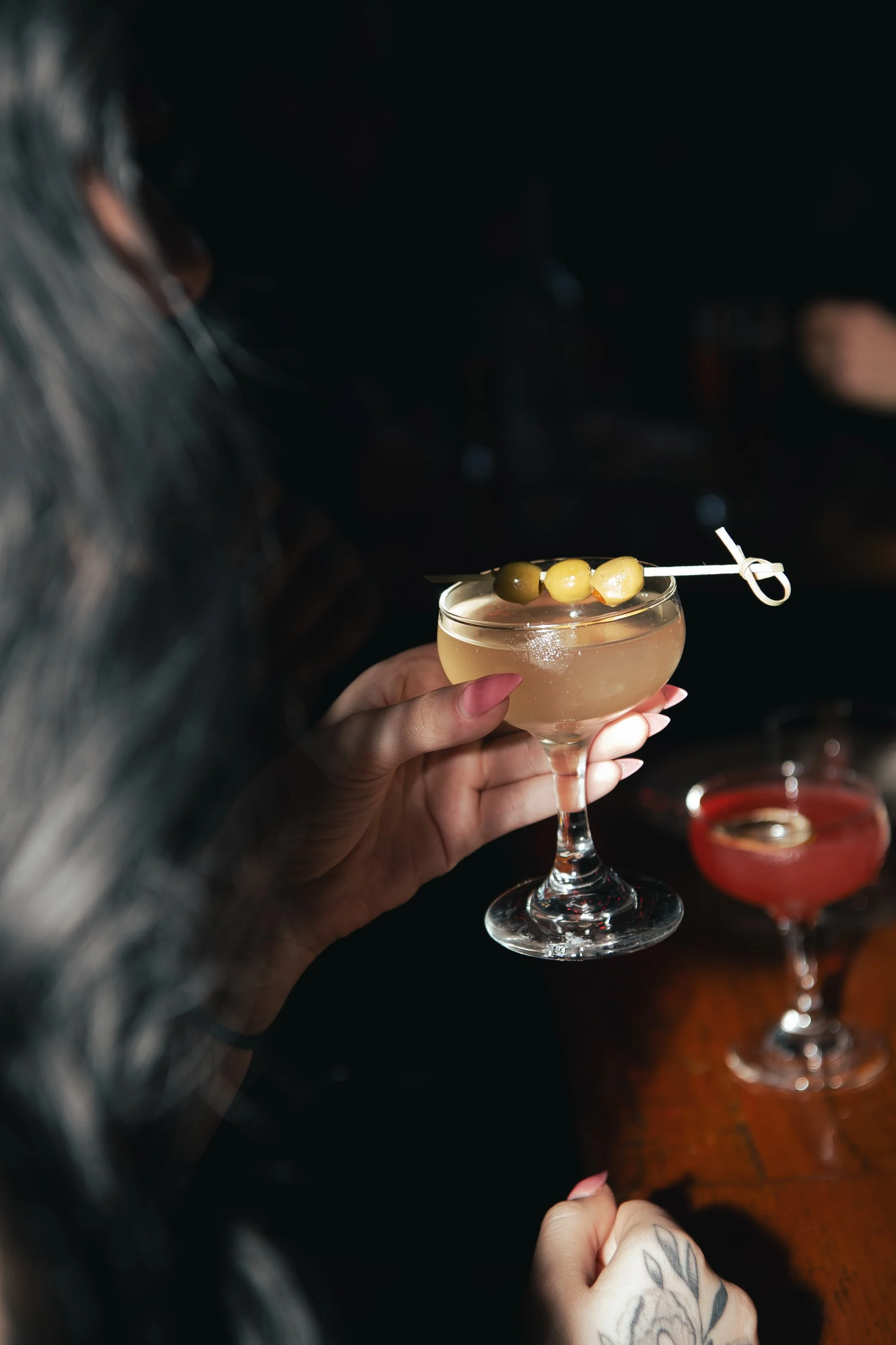 A creamy coffee cocktail topped with cocoa powder and coffee beans in a coupe glass, on a wooden board surrounded by scattered coffee beans and a small scoop of beans on a wooden surface.