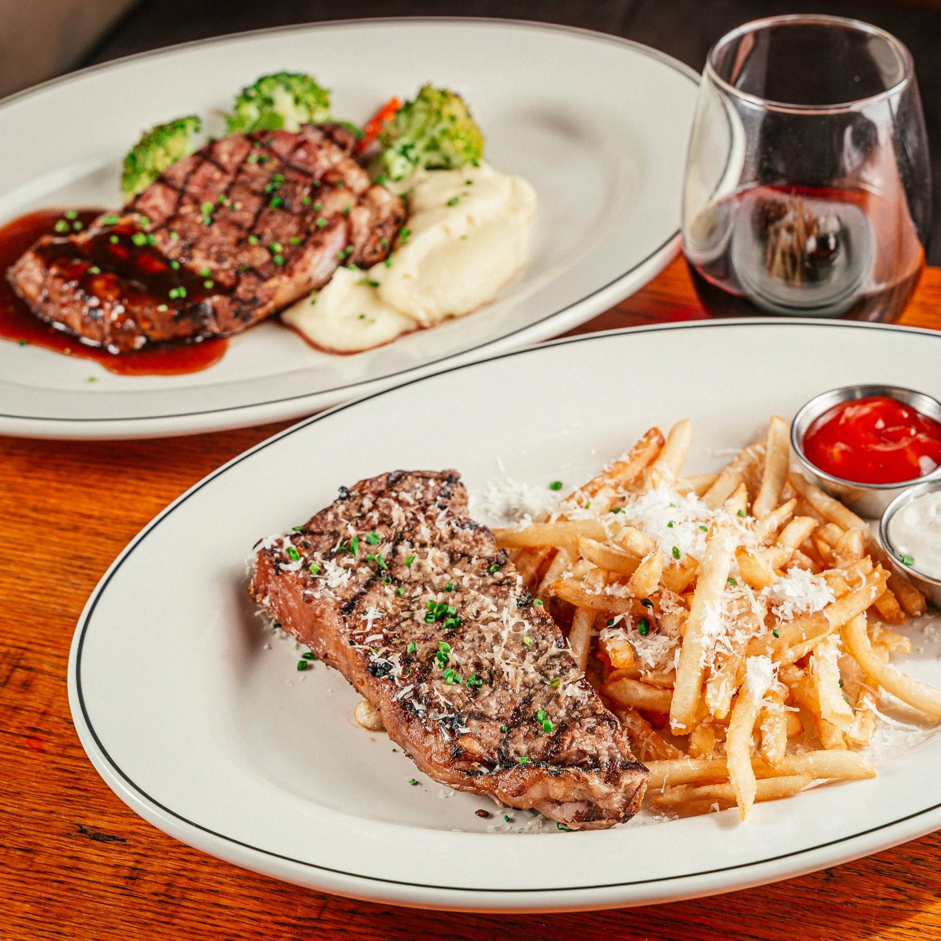Two plates of food on a wooden table, one with french fries topped with cheese and served with ketchup and ranch dips, and the other with a piece of grilled steak topped with herbs, and a second grilled meat dish with mashed potatoes, broccoli, and gravy in the background.