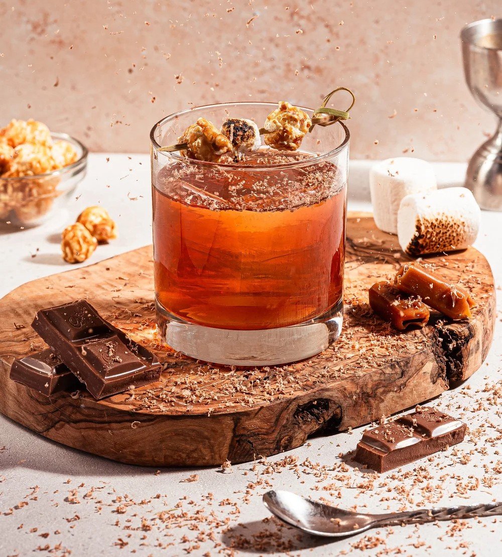 A caramel-garnished cocktail at Fox On John, surrounded by chocolate, toasted marshmallows, and caramel pieces.