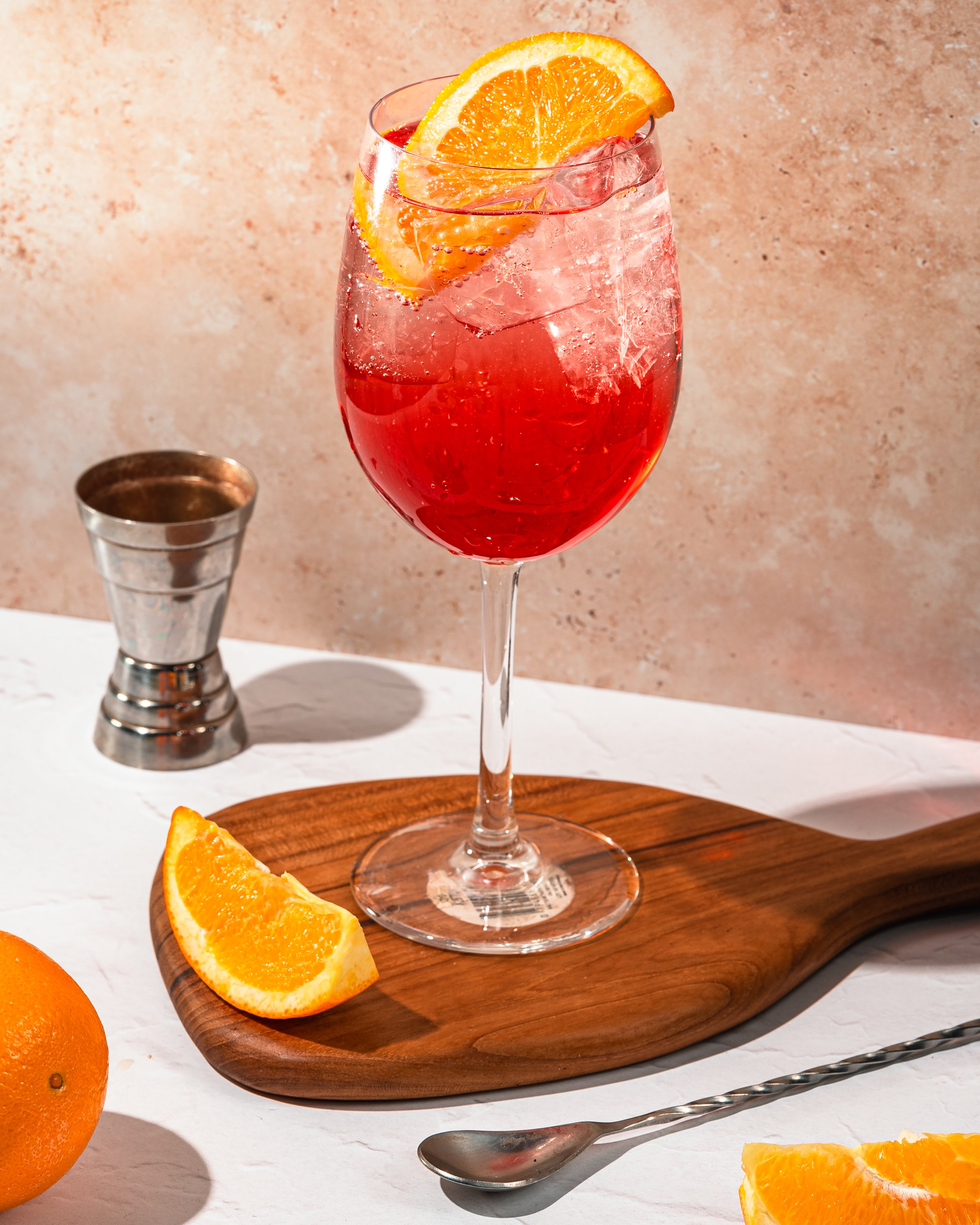 A glass of red beverage with ice and orange slices on top, resting on a wooden serving board. An orange is partially visible to the left, a metal jigger is in the background, and a spoon lies in front of the board.