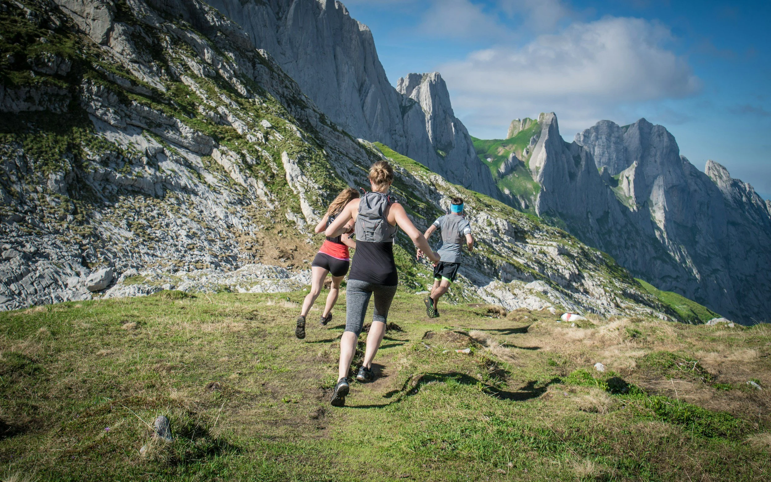 Group of people trail running on grassy mountain trail surrounded by rocky cliffs and scenic mountain landscape under blue sky.