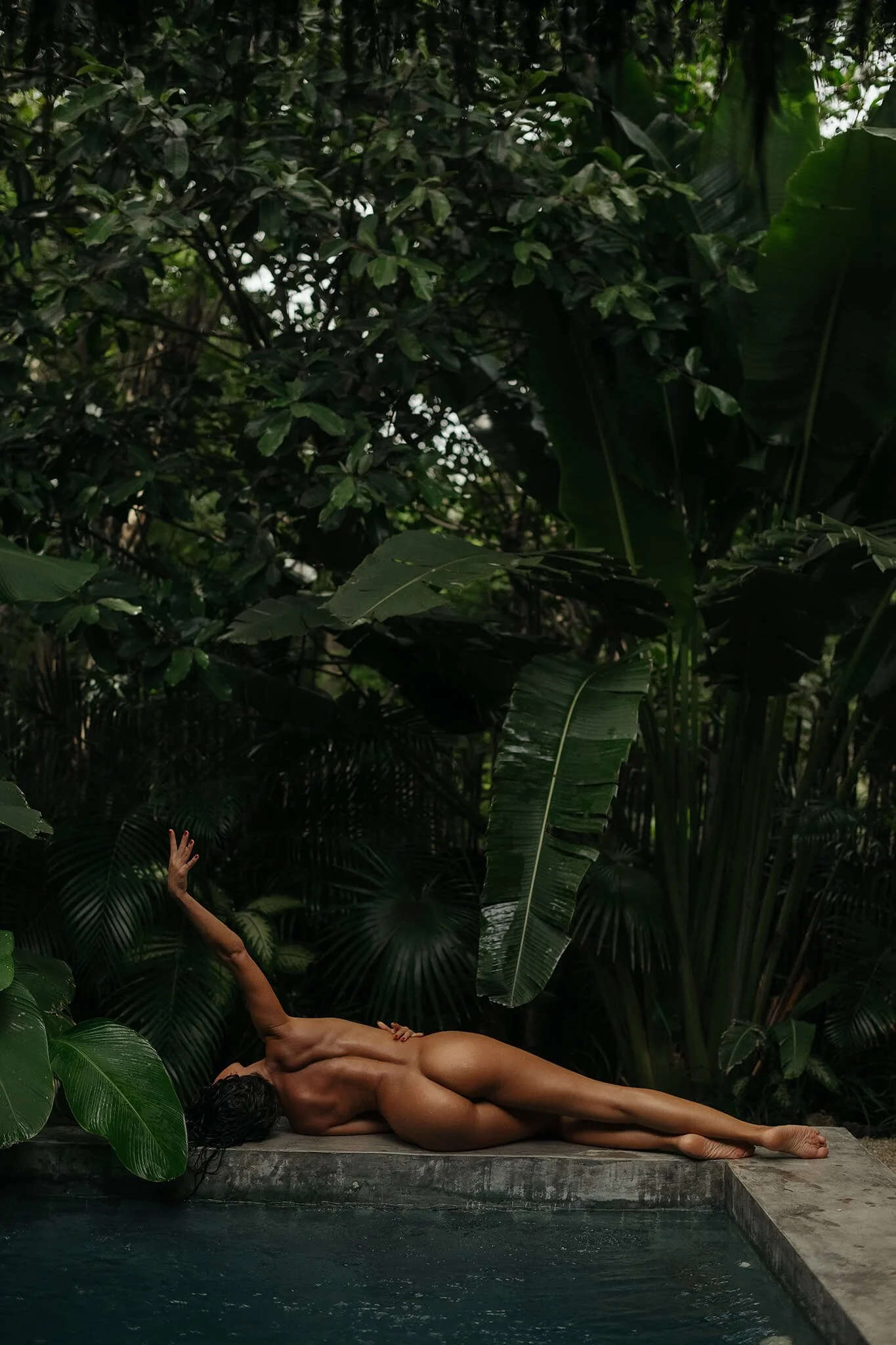 A nude woman lying on her side on a stone pool edge surrounded by lush green tropical plants, with her arm extended upwards.