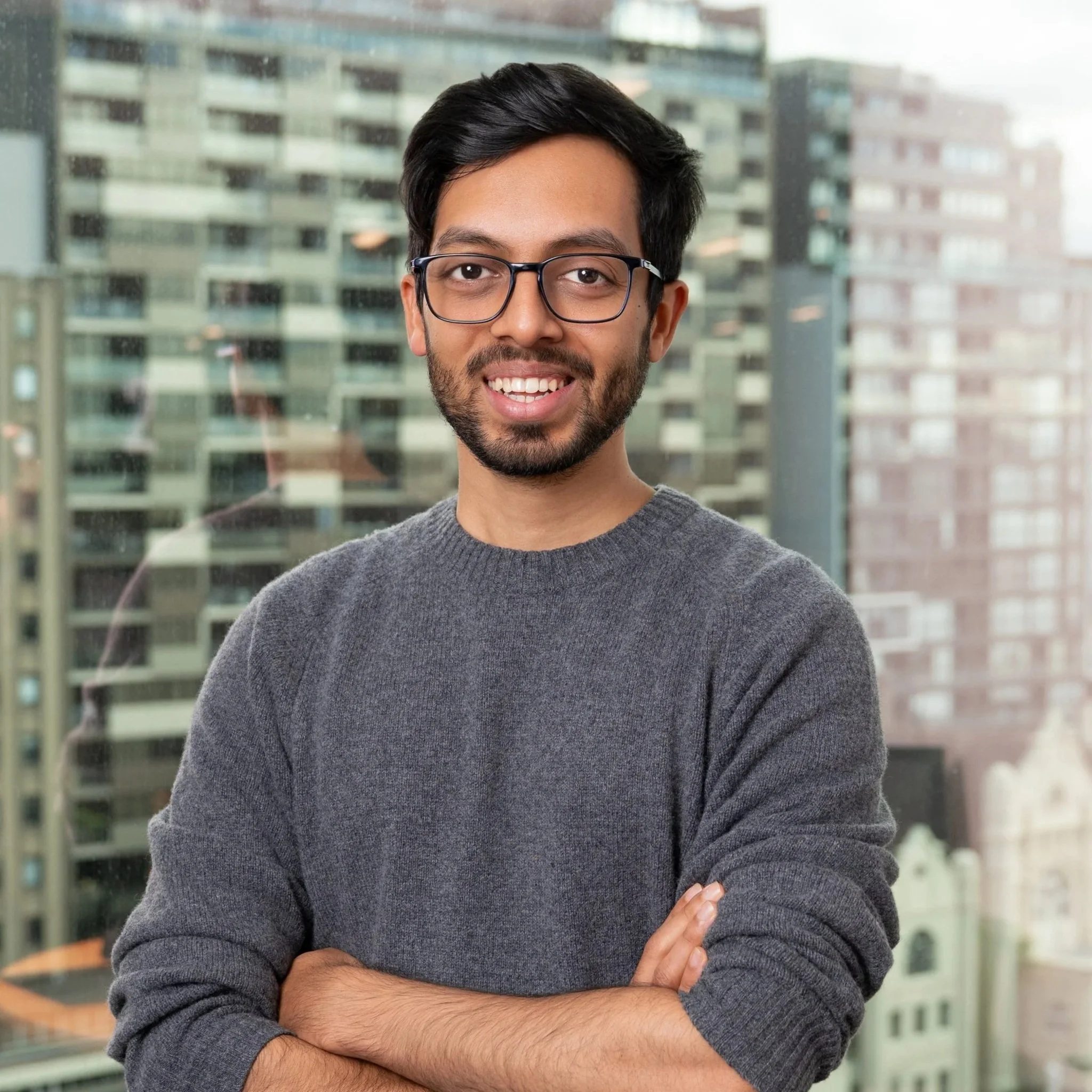 A man with short dark hair, glasses, and a beard, smiling, outdoors with green trees and a blue sky in the background, dressed in a suit jacket and light blue shirt.