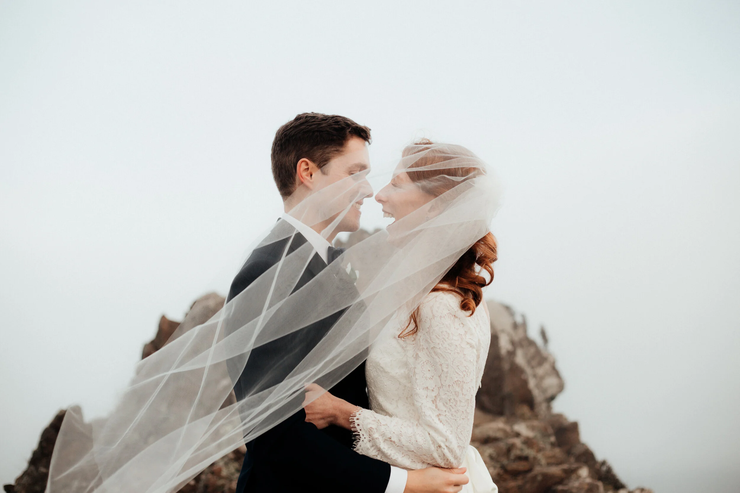 A bride and groom smiling and embracing outdoors with rocky terrain in the background.