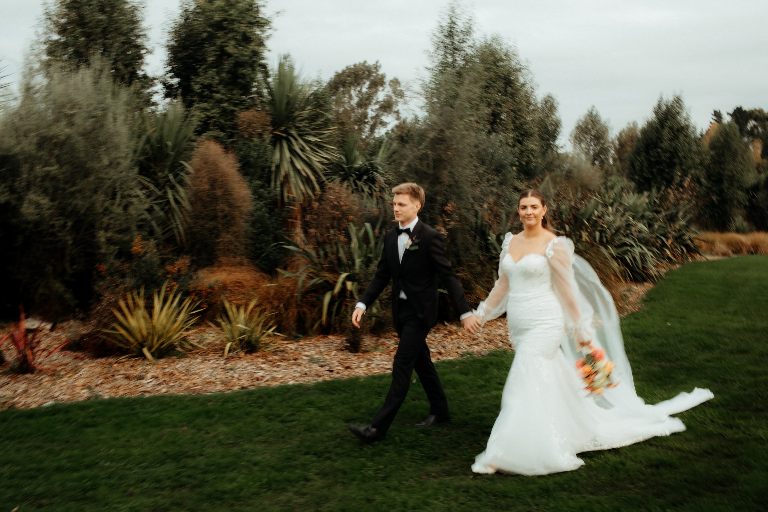 A bride and groom walking hand in hand outside on grass, surrounded by greenery and bushes. The bride is wearing a white wedding dress with long tulle sleeves and holding a bouquet, while the groom is dressed in a black tuxedo with a bow tie.