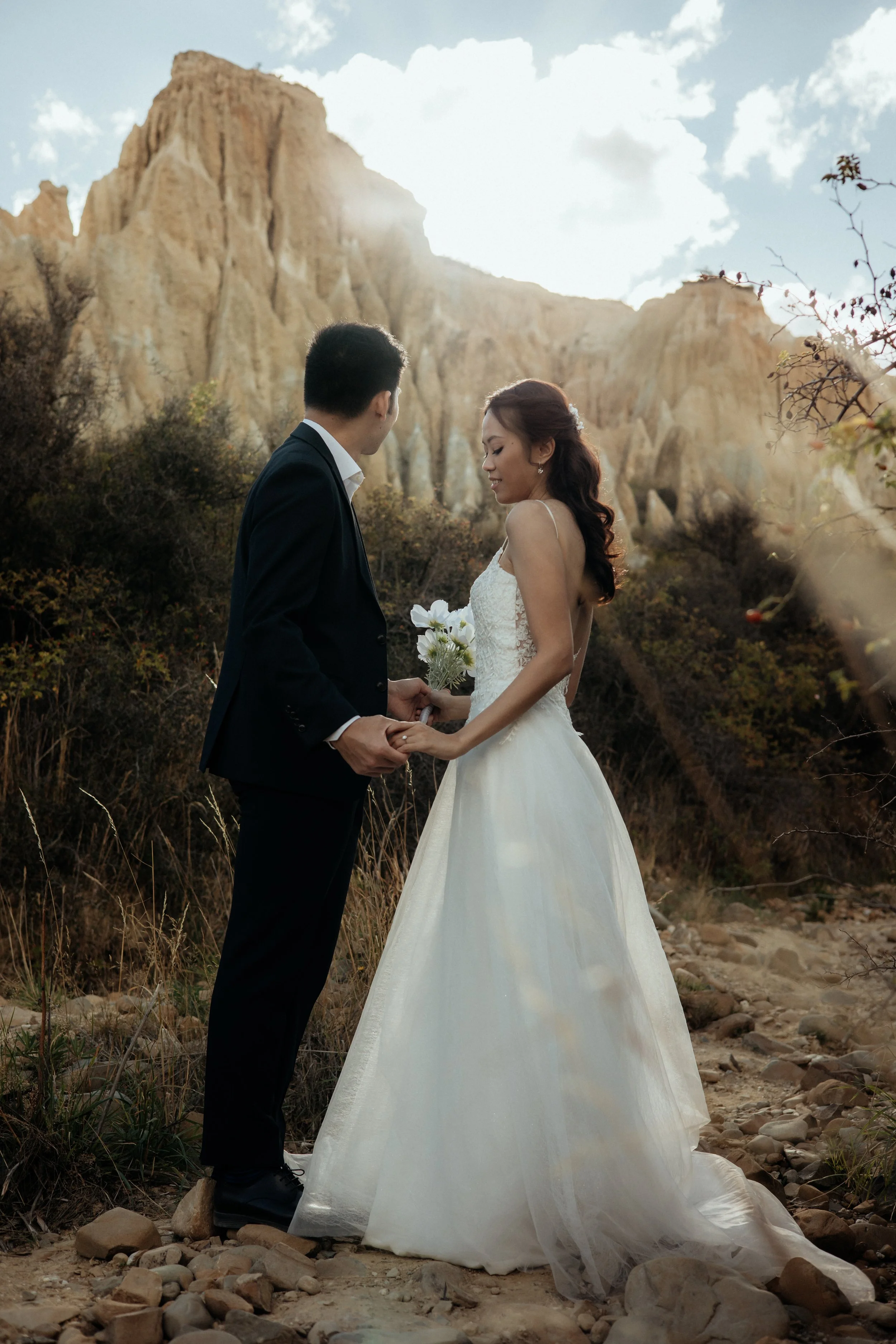 A bride and groom holding hands outdoors, standing on rocky terrain with a mountain in the background, during sunset.
