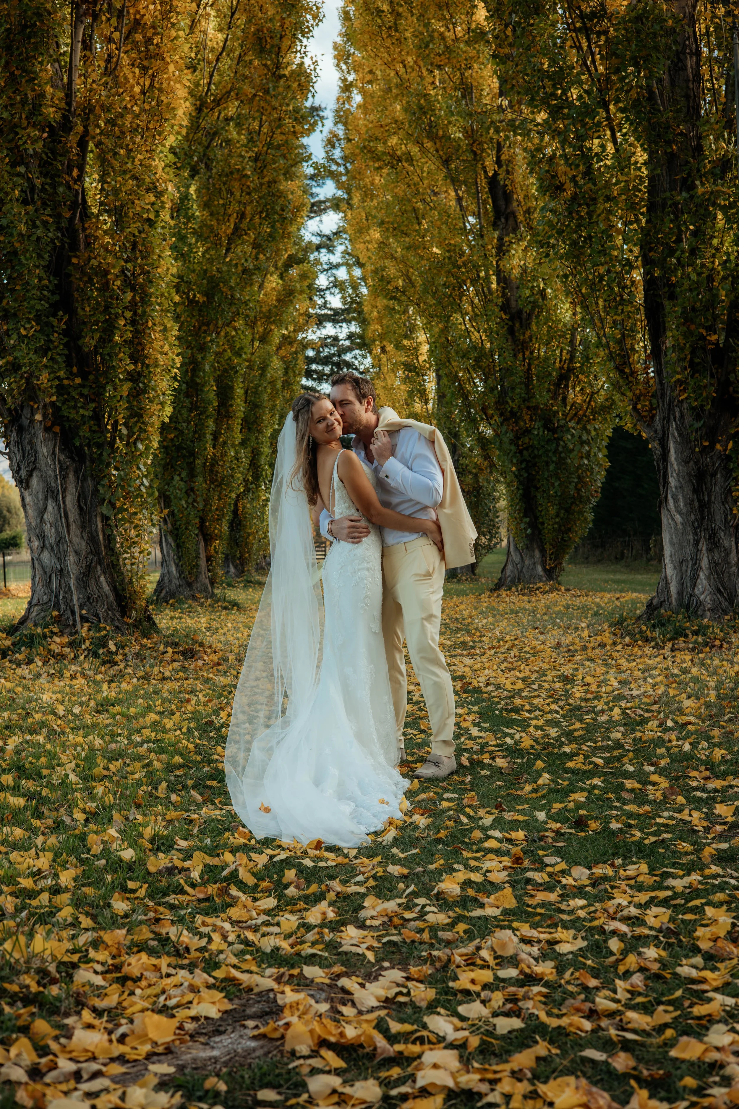A newlywed couple in wedding attire sharing an embrace on a path covered with fallen autumn leaves, lined with tall trees with green and yellow foliage under a clear sky.