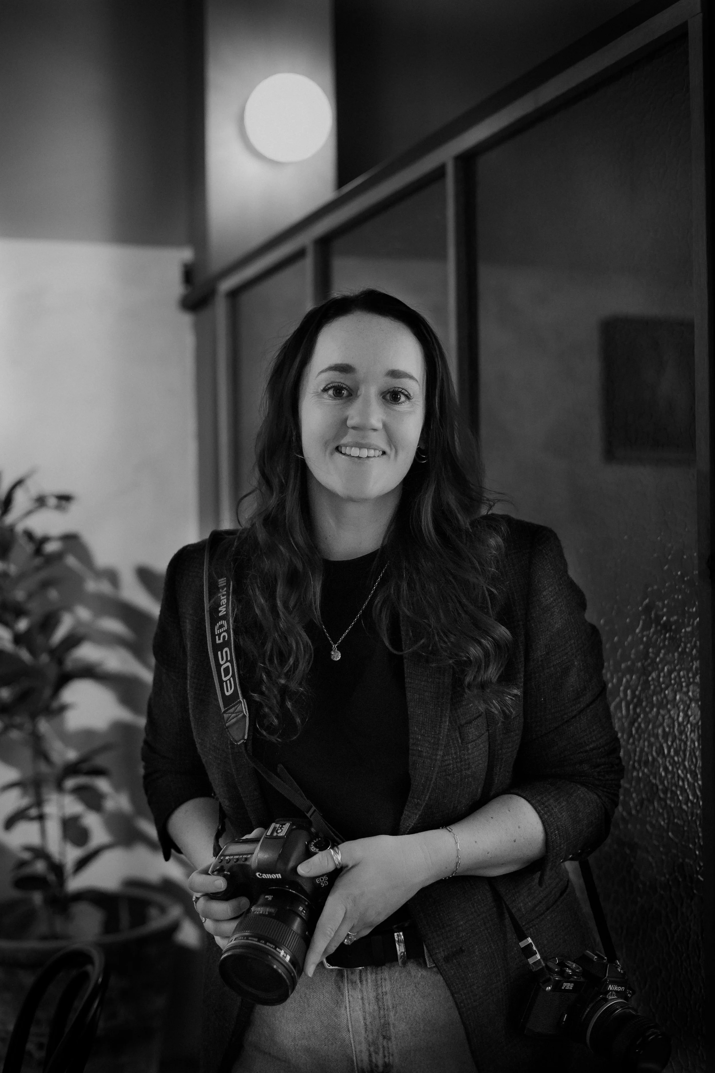 A smiling woman holding a Canon DSLR camera, standing indoors near a textured wall and a potted plant.
