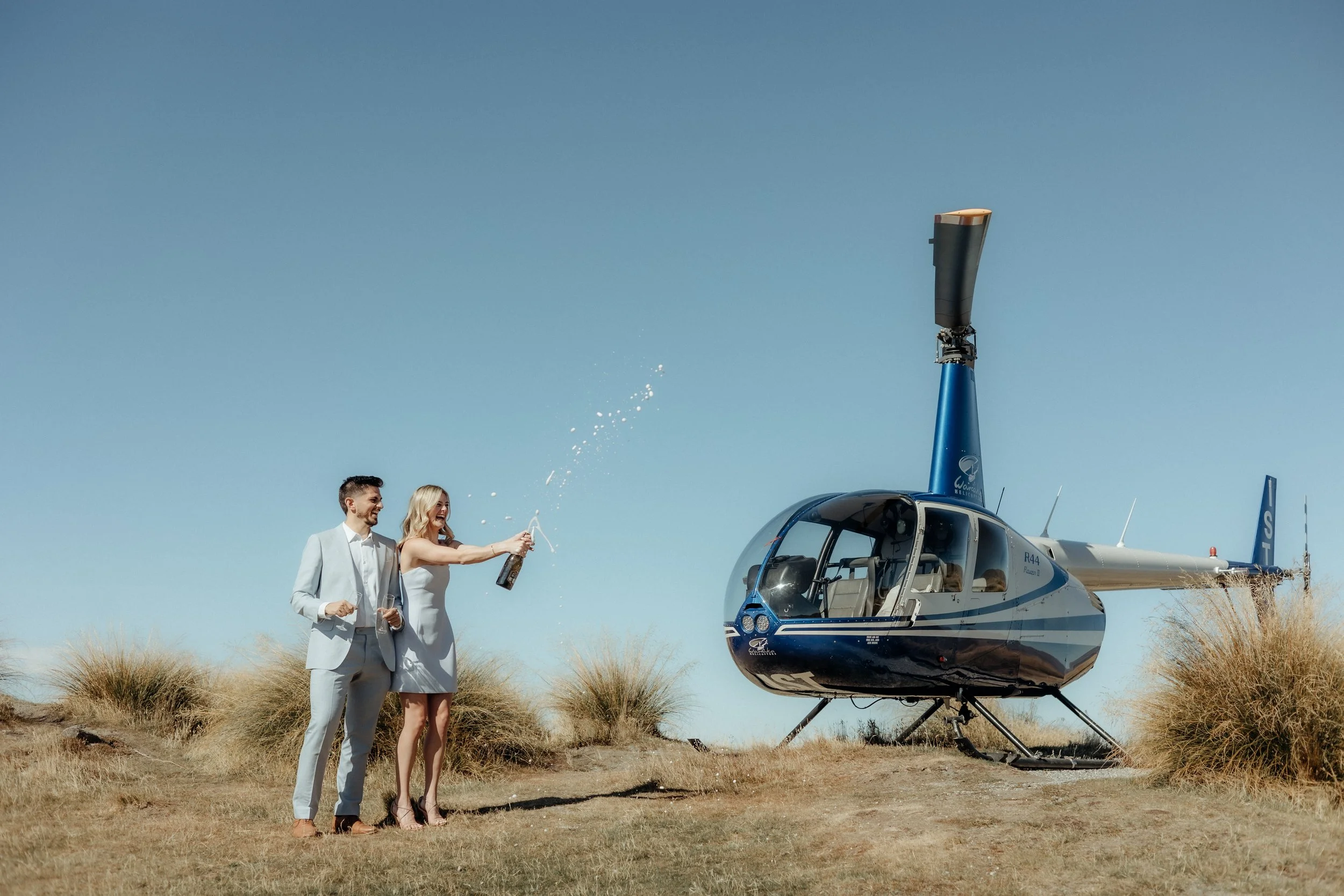 A couple in wedding attire celebrating outdoors near a helicopter, with the woman opening a bottle of champagne, and the man smiling, on a clear day with tall grass.