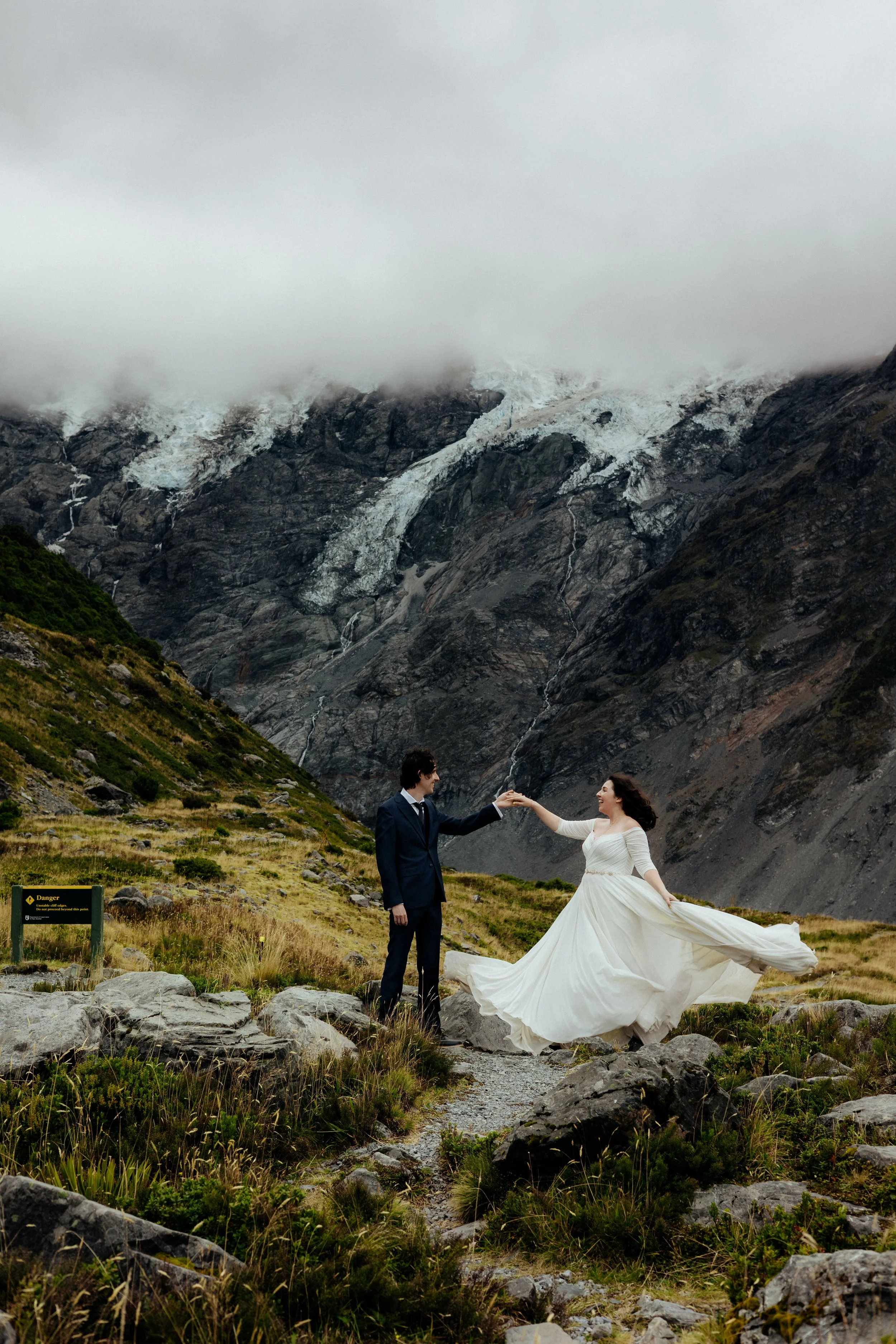 A couple in wedding attire dancing on a rocky trail in a mountainous landscape with snow-capped peaks and a cloudy sky.