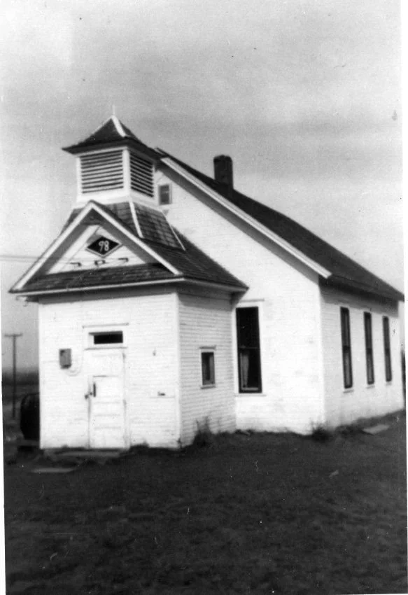 A black and white photo of a simple school house. There is a "98" displayed above the door and a bell tower above that.