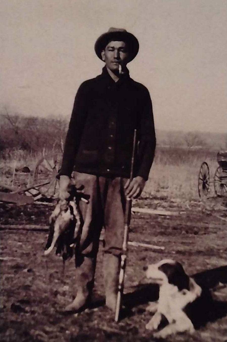 A black and white photo of a tall man in a button down shirt, jeans, and boots. He is wearing a cowboy hat and holding a hunting rifle in one hand and an unknown animal in the other. A border collie dog lies at his feet