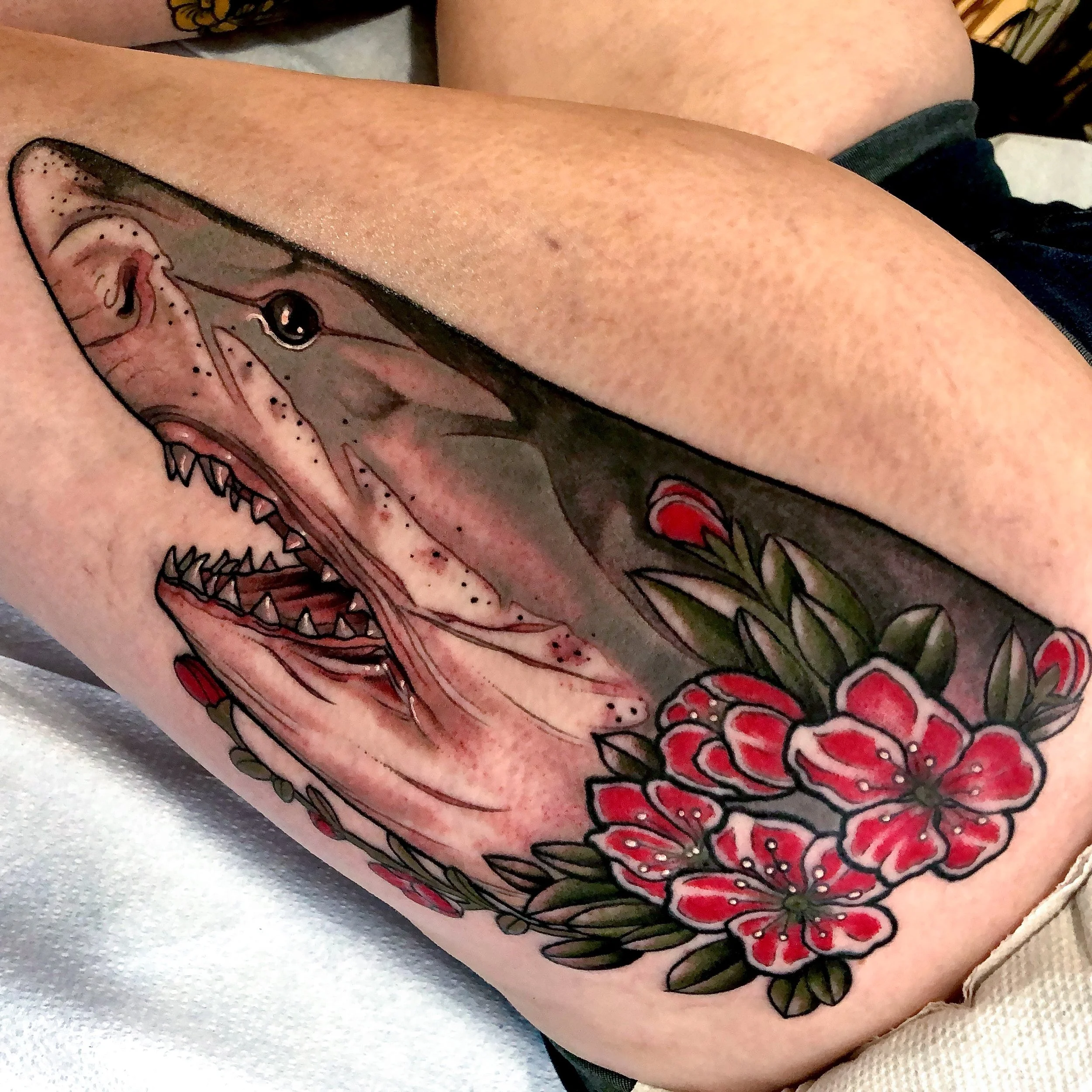 Close up photo of a tattoo on the upper thigh of a great white shark head, it is shaded gray and white, and is framed with red and white flowers.