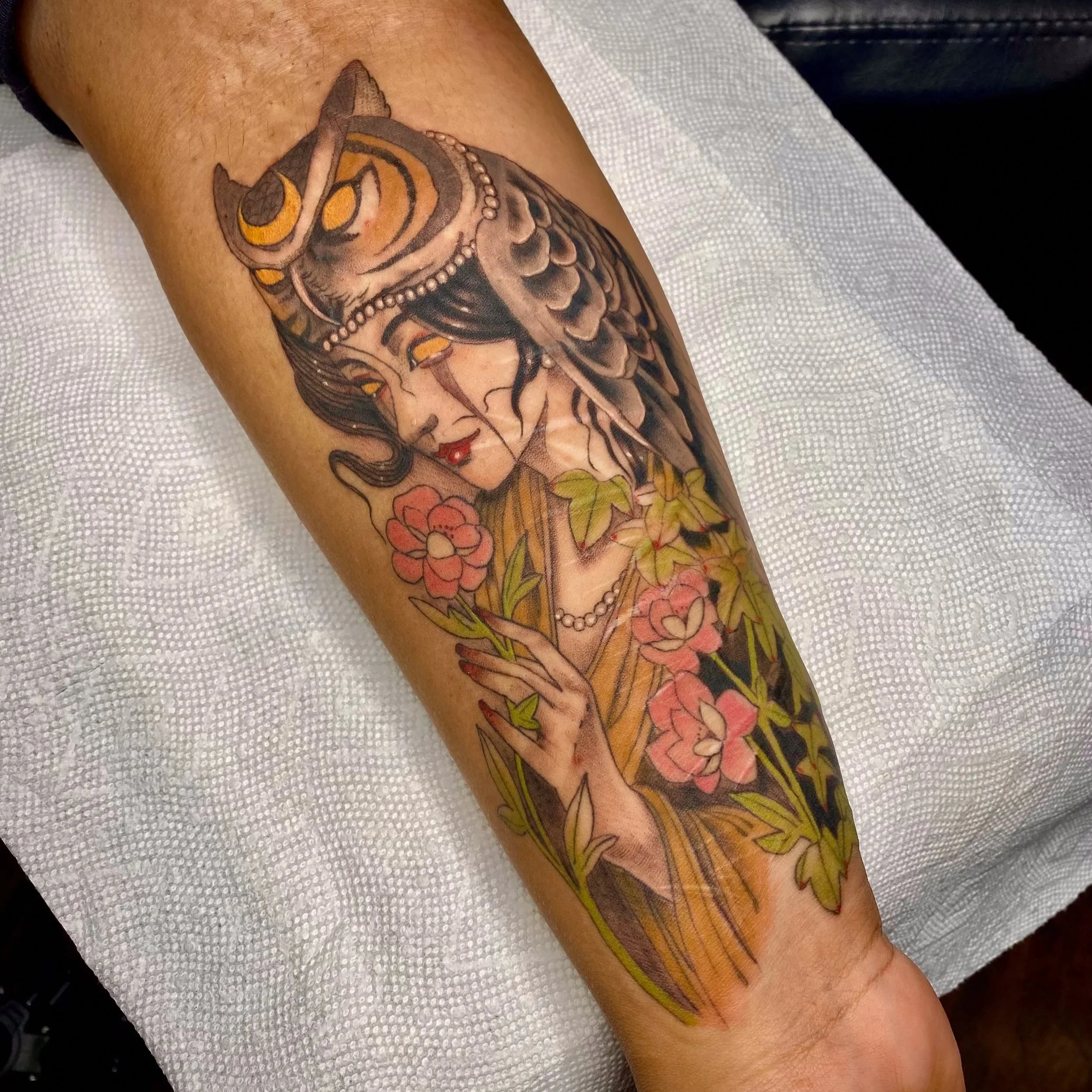 A photo of a tattoo on the wrist that is covering very faint scarring. It is a portrait of a woman with an owl headdress adorning her. She is holding a pink flower and crying.