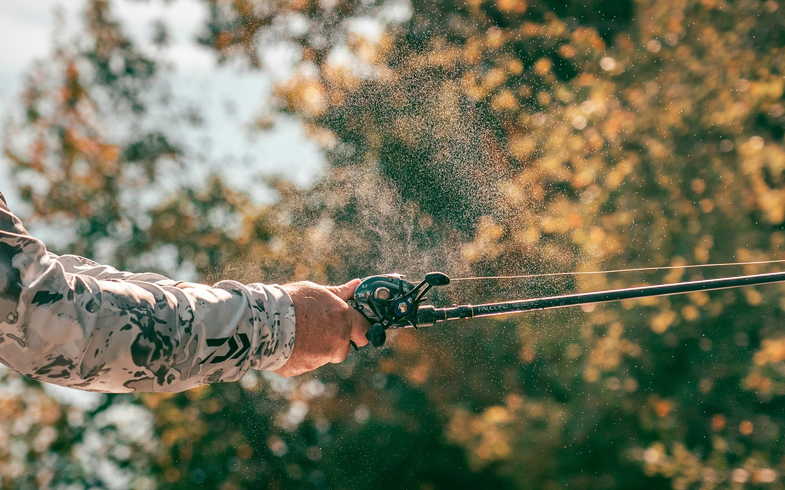 A person wearing a long sleeve camouflage shirt is fishing with a reel and rod, casting a line into the water, with sunlight filtering through trees in the background.