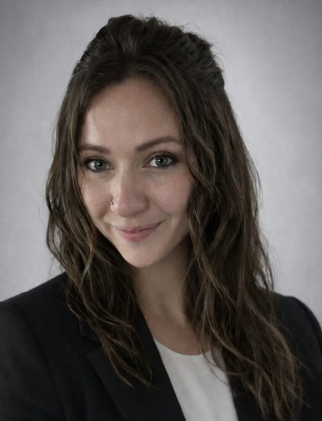 Portrait of a woman with wavy brown hair and blue eyes, wearing a black blazer and white top, smiling softly at the camera with a neutral background.