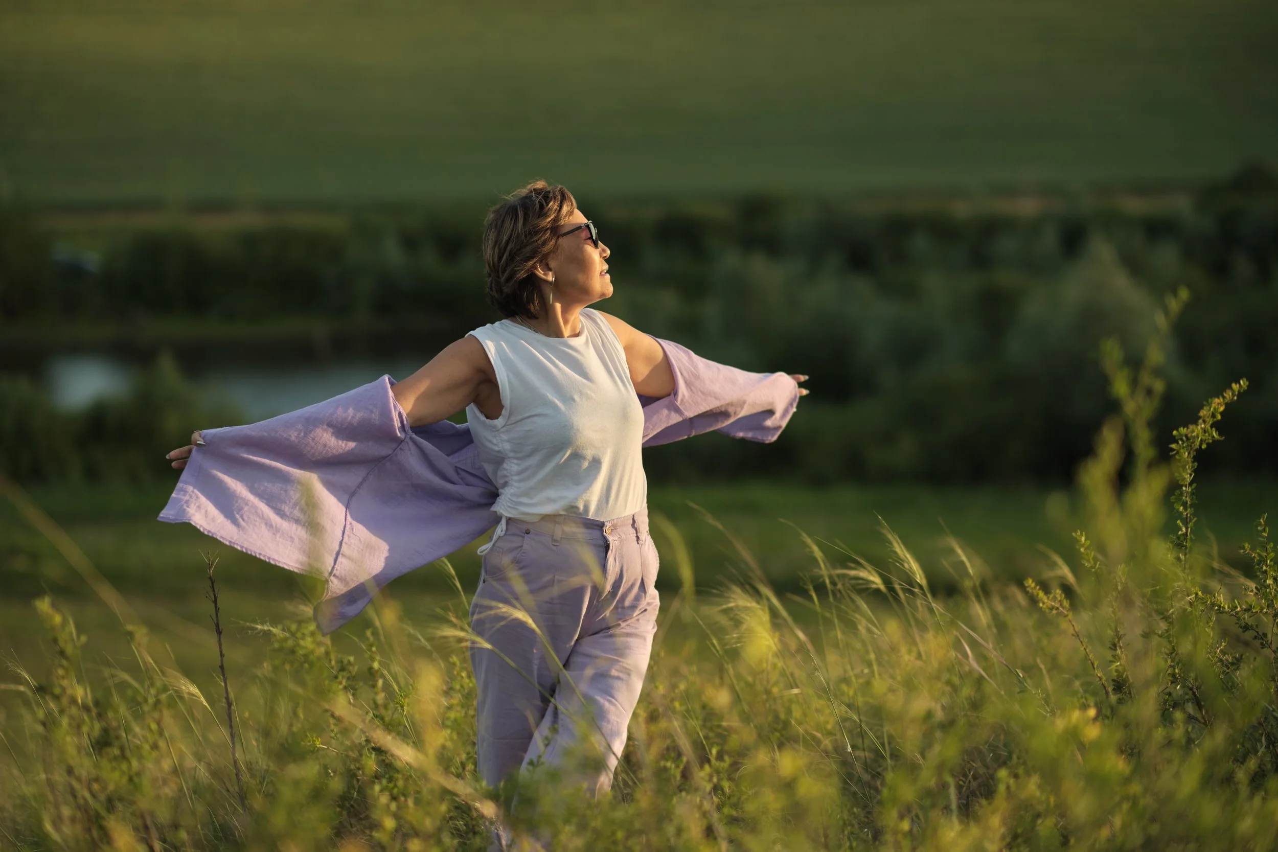 woman standing in an open field with arms wide, representing deep core support, alignment and long term pain prevention.