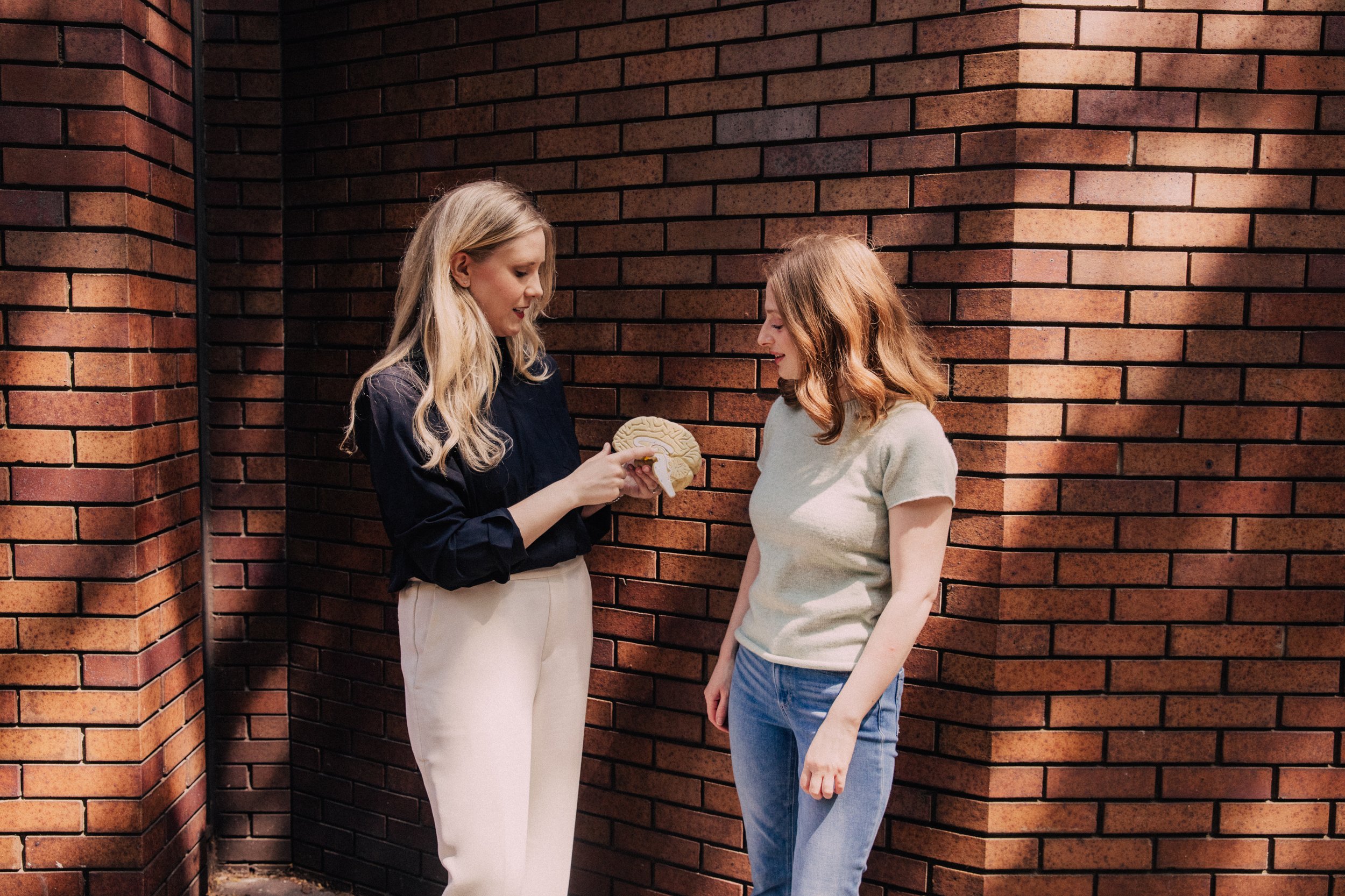 Two women standing against a brick wall, one showing a model of a brain to the other. Represents the team of experts behind New York Women's CBT, offering niched online therapy in New York for women with chronic illness and chronic pain.