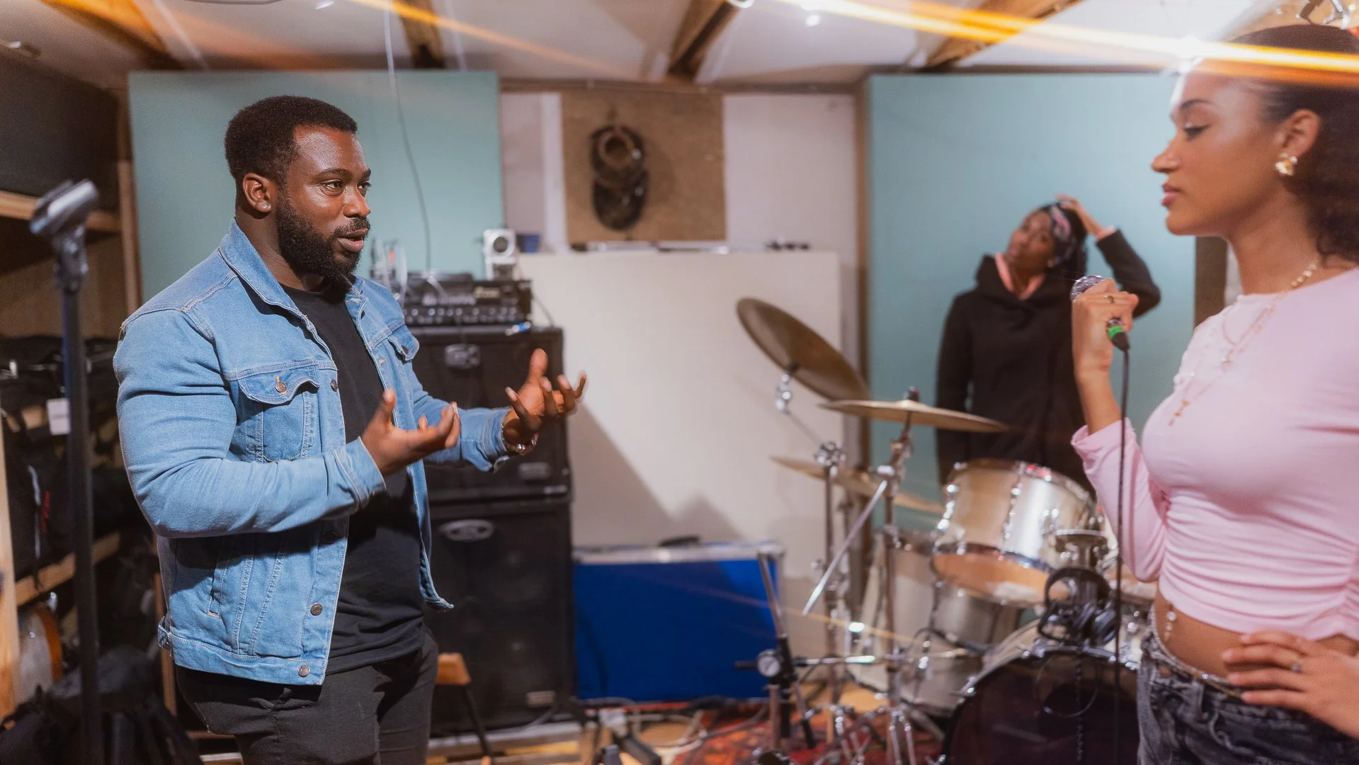 A man in a denim jacket is speaking or explaining while a woman in a pink top holds a microphone in a music rehearsal studio with drums and sound equipment in the background.