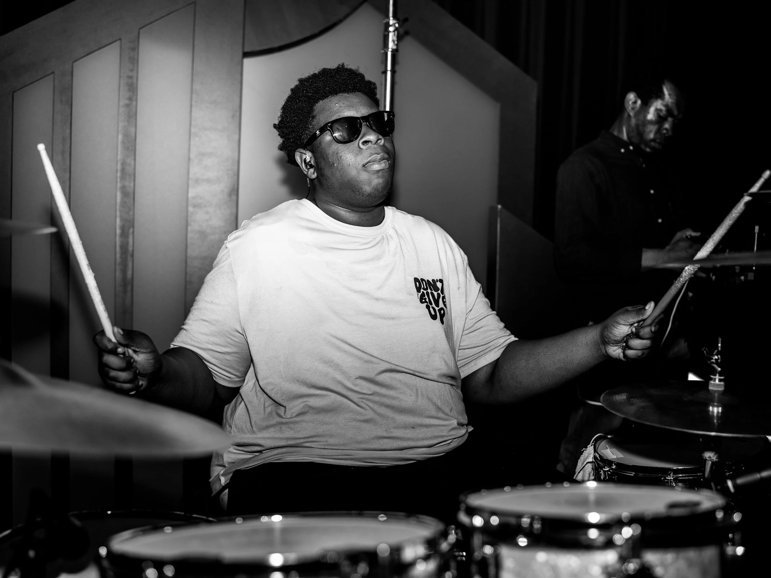A Black man wearing sunglasses and earphones sits behind a drum kit, holding drumsticks, in a music studio with acoustic panels on the wall.