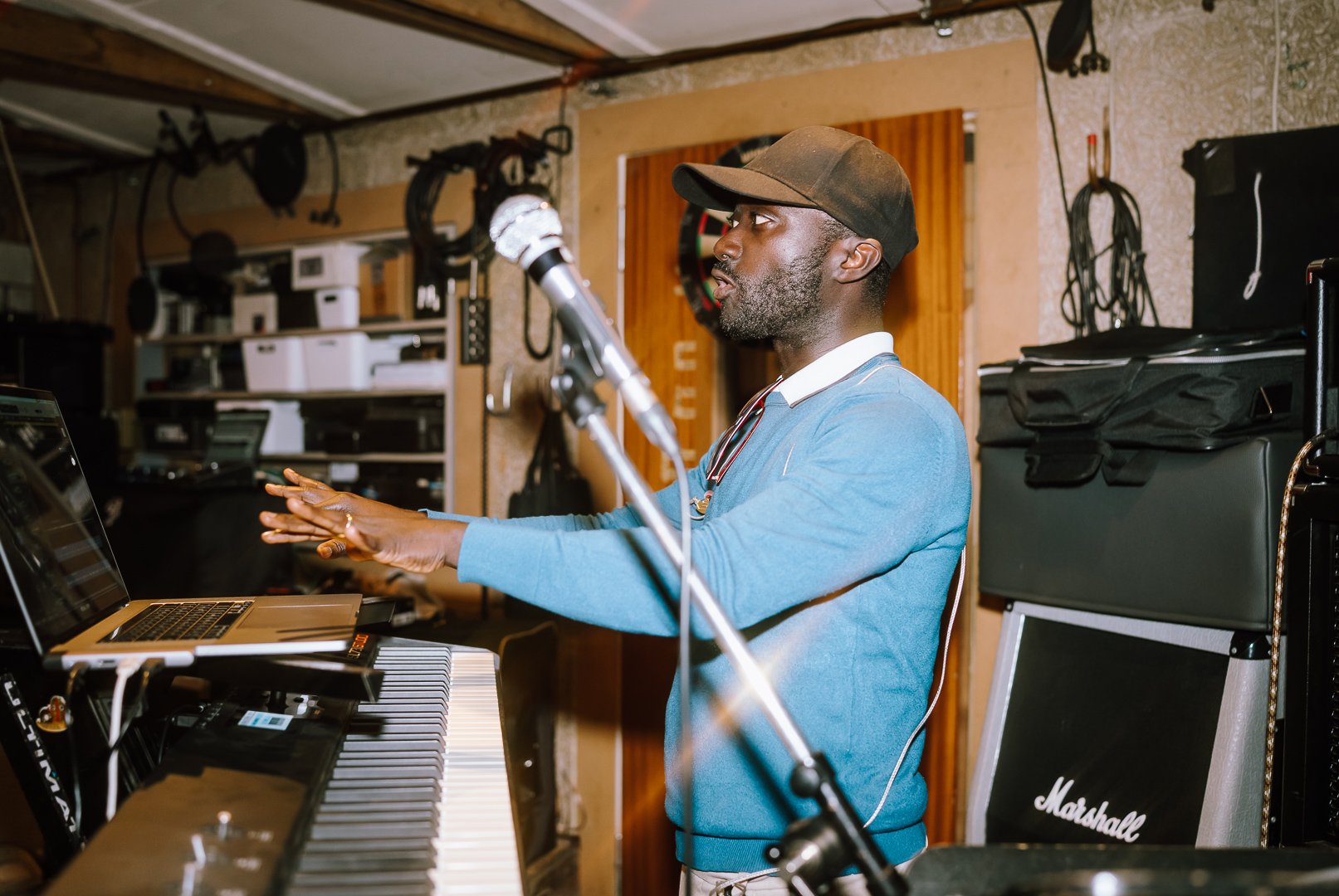 Obed Owusu wearing a hat and blue sweater playing a keyboard in a music studio, with various equipment and a microphone in front of him.
