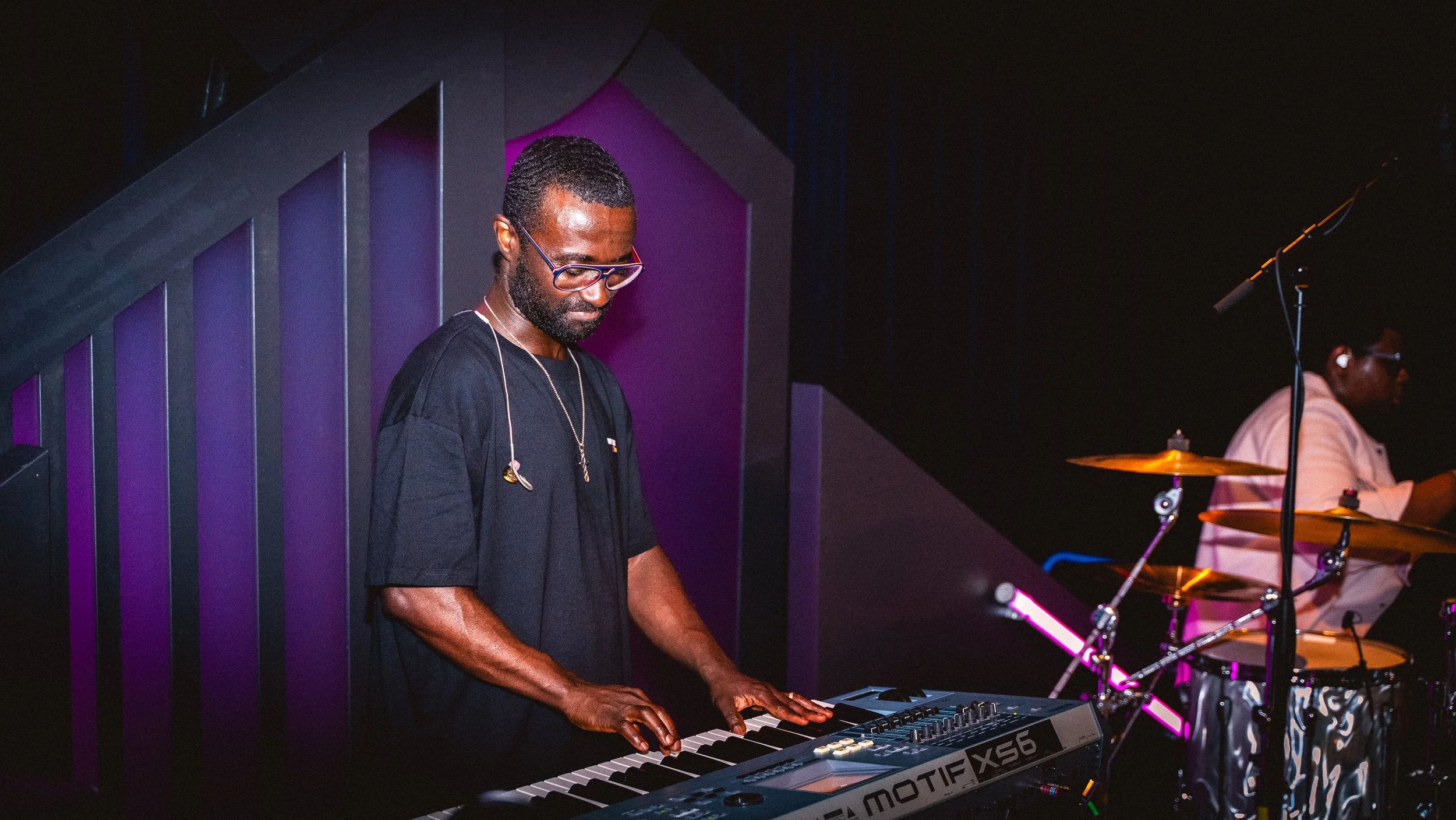 Obed Owusu is playing the keyboard, with Justice in the background on drums, in a dimly lit, purple-hued music venue.
