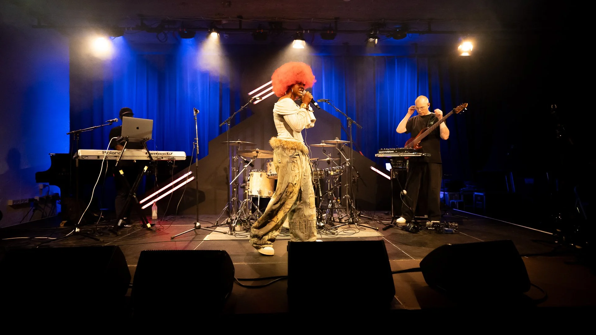 A band is performing on stage with a female singer, Mary Jane, with large pink curly hair, The stage has blue curtains and stage lights above.