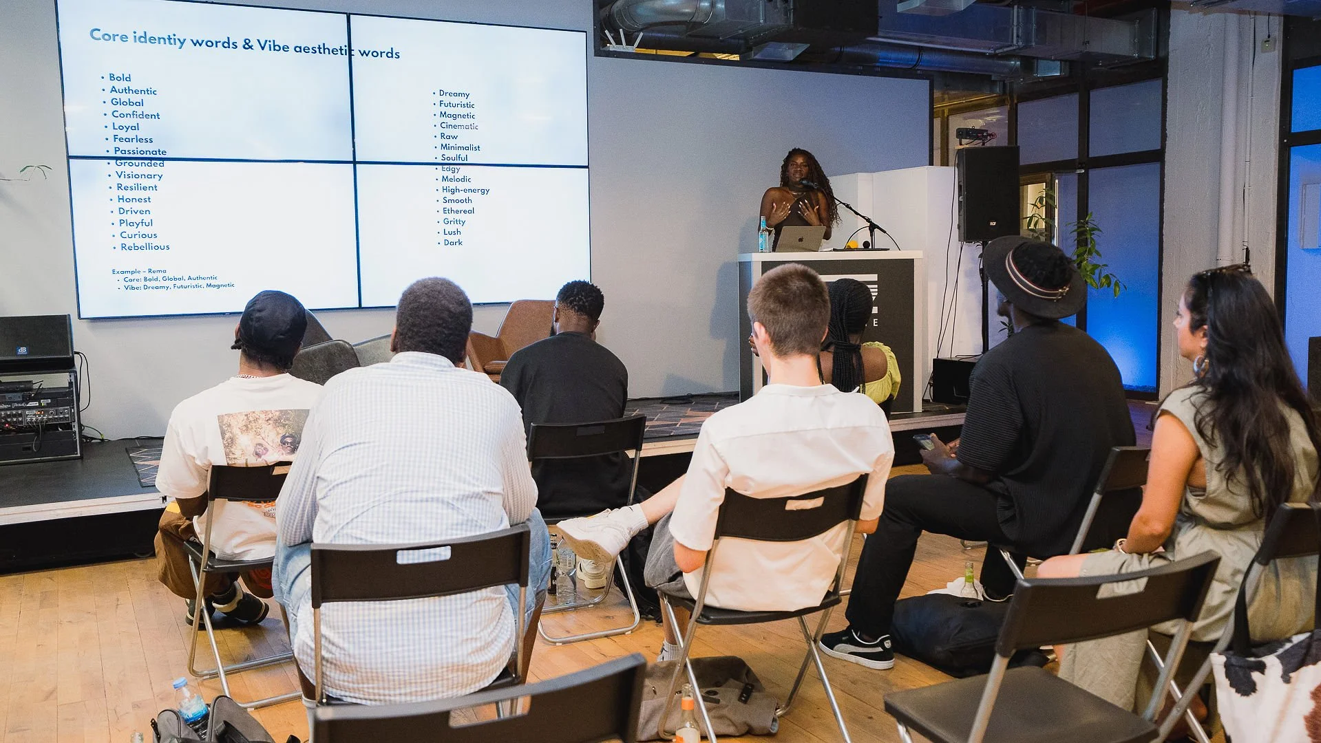 A woman with curly hair giving a presentation to an audience in a modern, industrial-style room. The presentation slide on the large screen displays words related to core identity and vibe aesthetics. The audience is sitting on black chairs, some wit