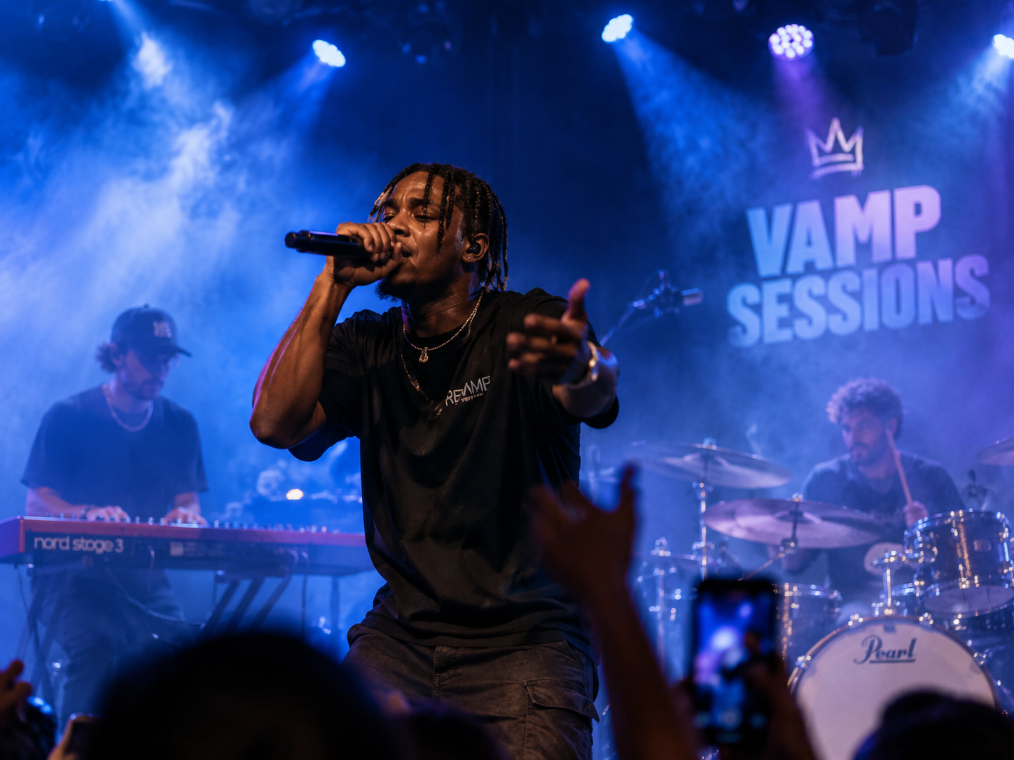 A rapper performs on stage during a concert with a band behind him. The stage has a sign that reads 'VAMP SESSIONS' with a crown logo above it. The rapper is holding a microphone and wearing a black t-shirt with the logo. Two musicians play keyboard and drums in the background, and audience members are visible in the foreground, some holding up phones.